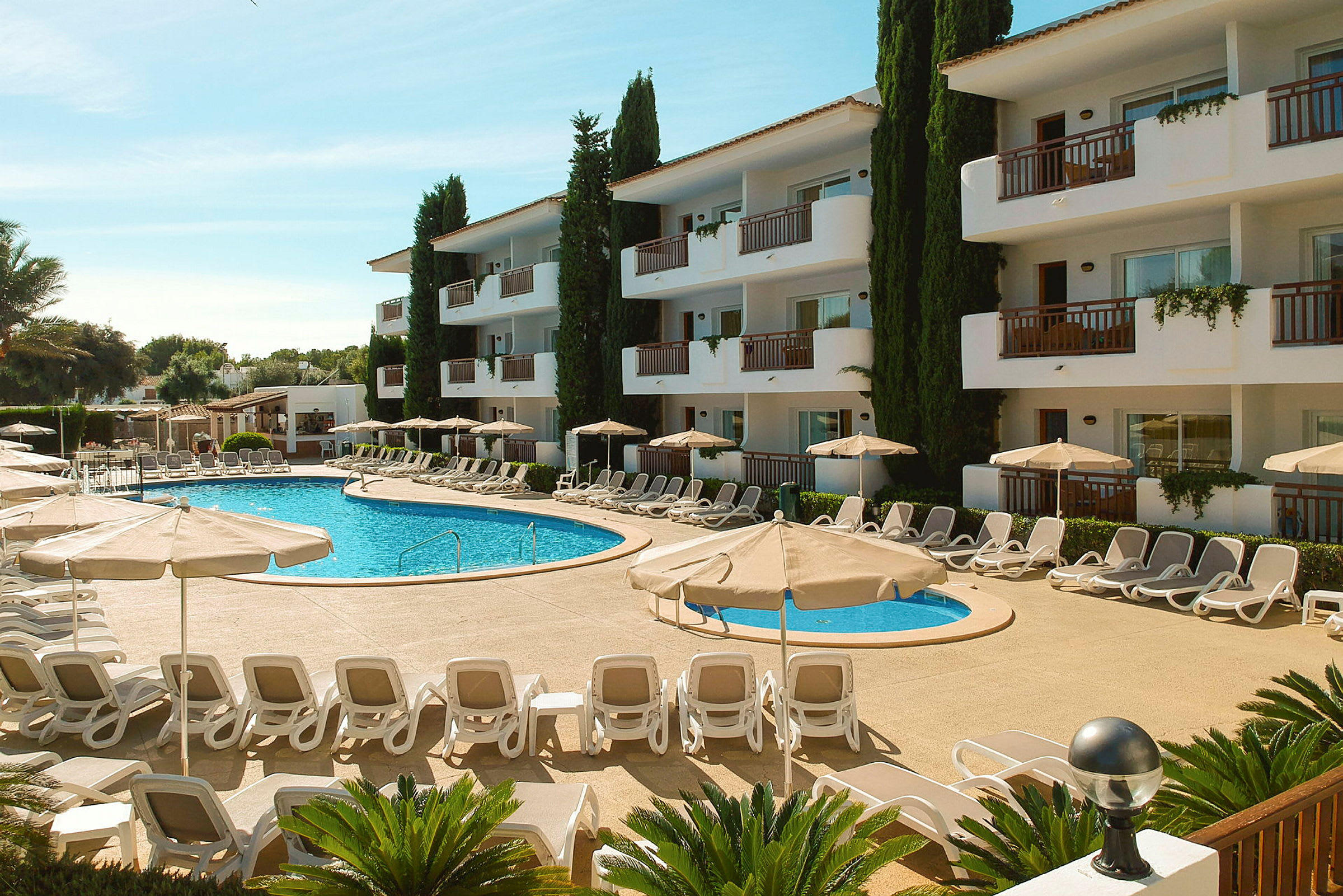 Bild av en hotellpool omgiven av solstolar och parasoller, med en vit byggnad i tre våningar i bakgrunden. (AI-genererad text)