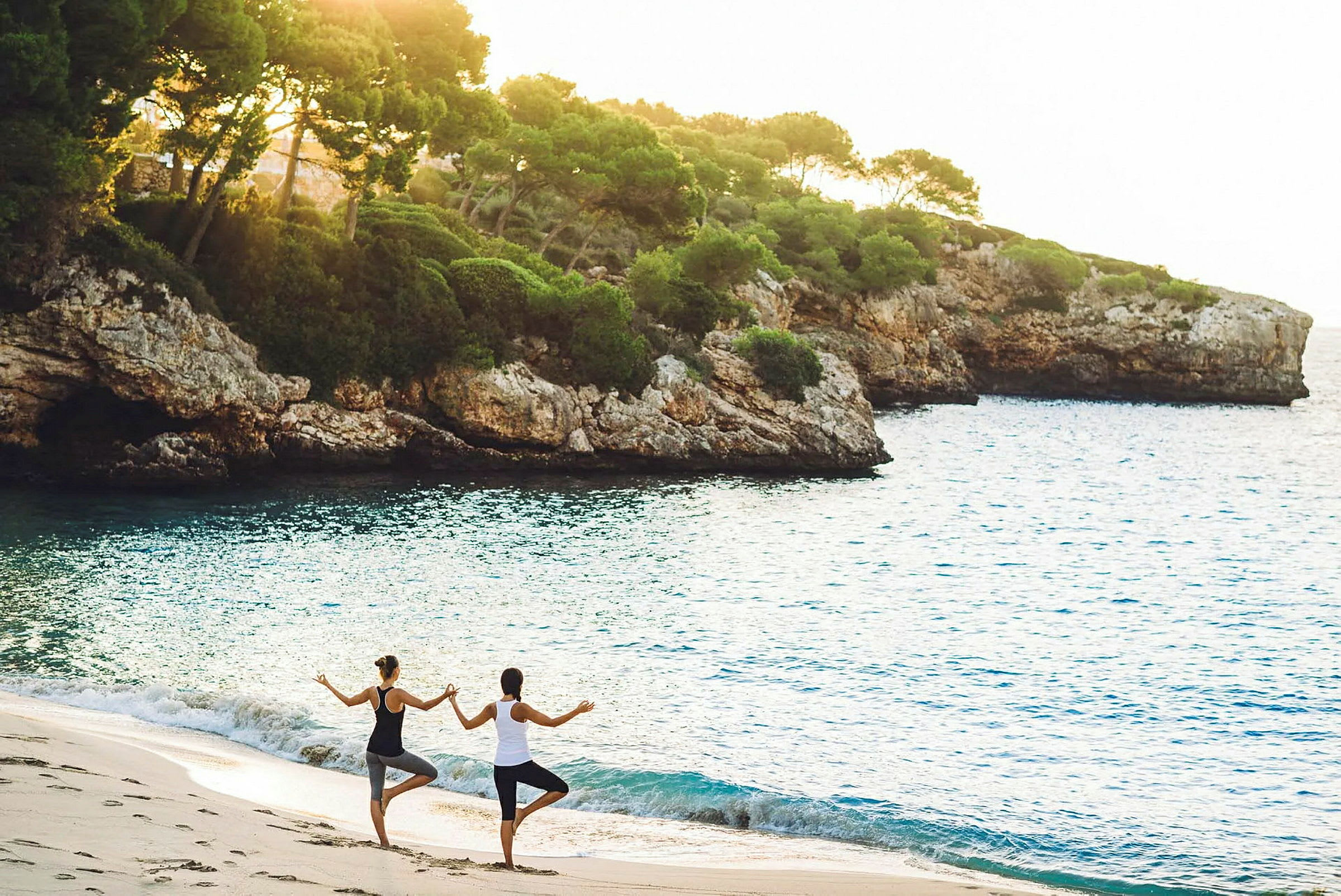 Två personer utövar yogapositioner på en sandstrand framför klart vatten och klippor. (AI-genererad text)