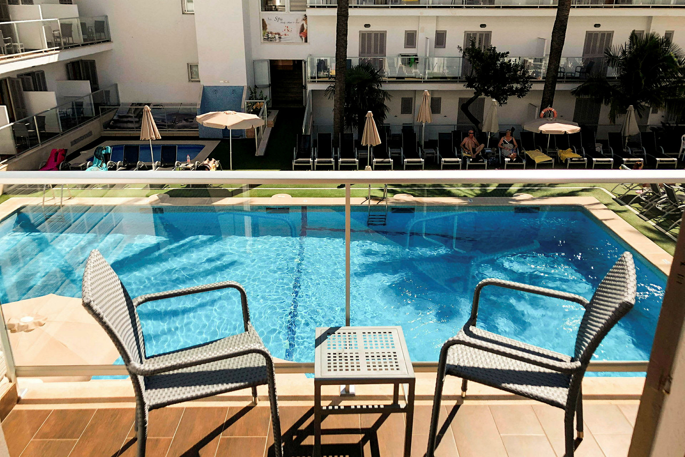 En pool med solstolar och parasoller vid ett semesterhotell, sedd från en balkong med stolar och ett bord. (AI-genererad text)