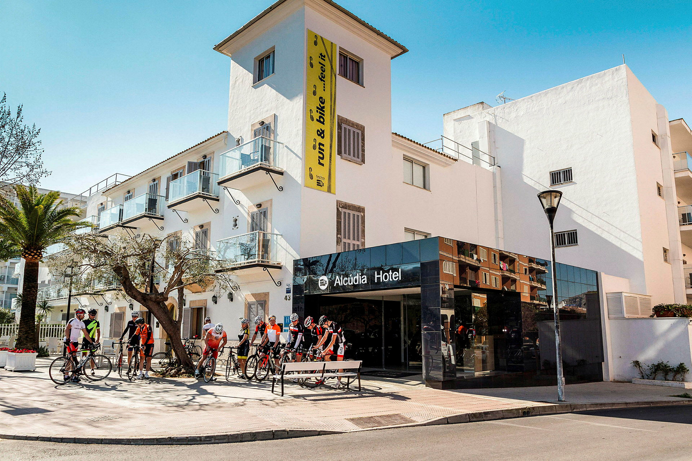 Vy av Alcudia Hotel med cyklister samlade framför ingången. (AI-genererad text)