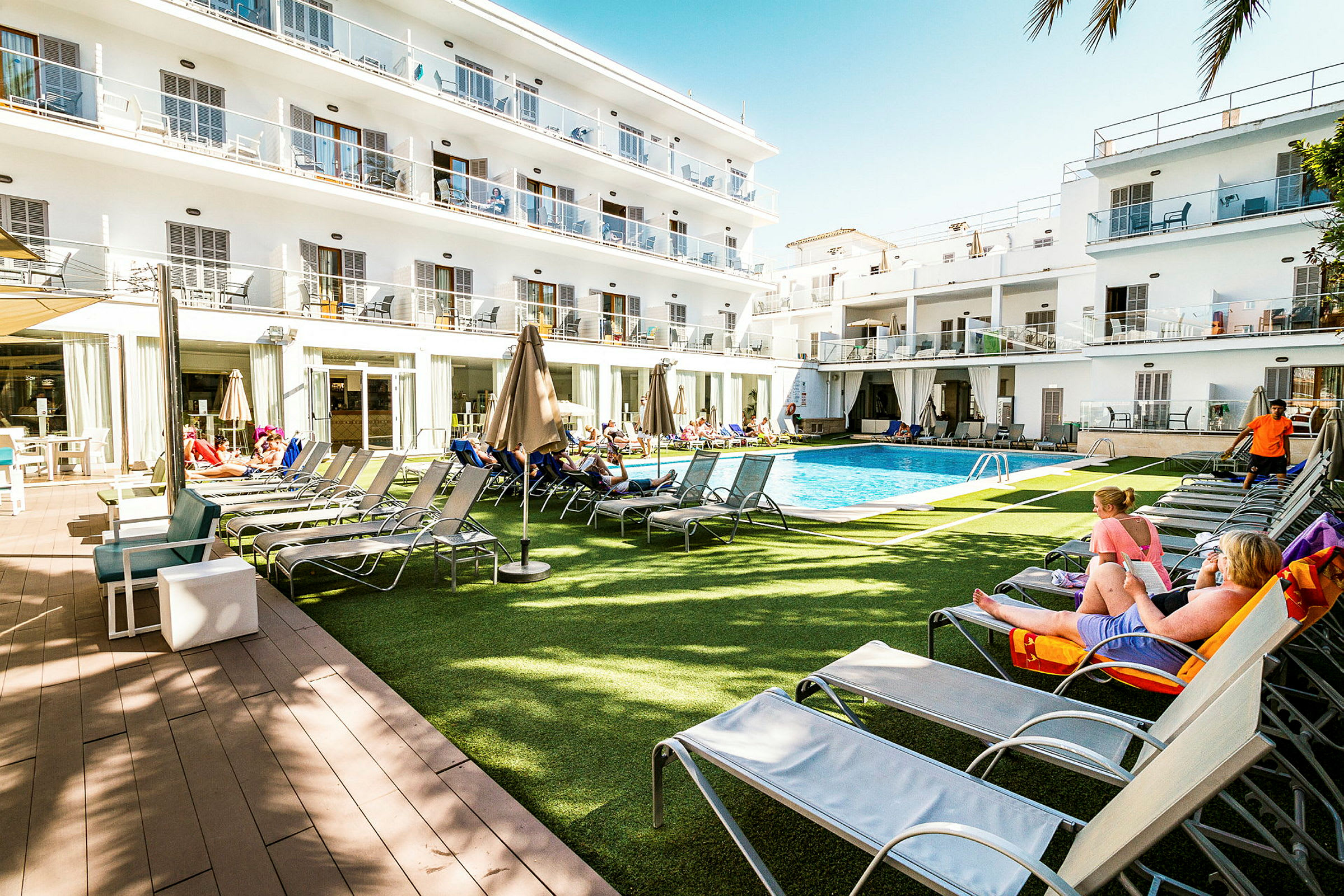 Lägenhetshotellområde med pool, solstolar och parasoller omgivna av vita byggnader med balkonger. (AI-genererad text)