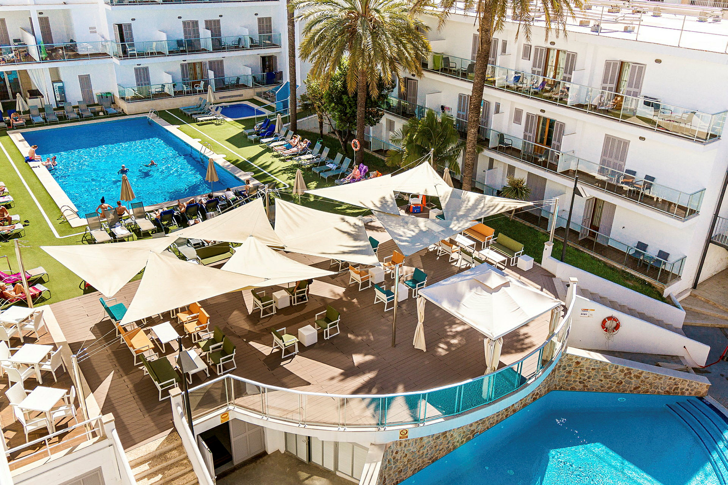 En bild av en hotellresort med en större pool omgiven av solstolar och parasoller. I förgrunden finns en utomhusbar med mängder av sittplatser under skuggande segeldukar. (AI-genererad text)
