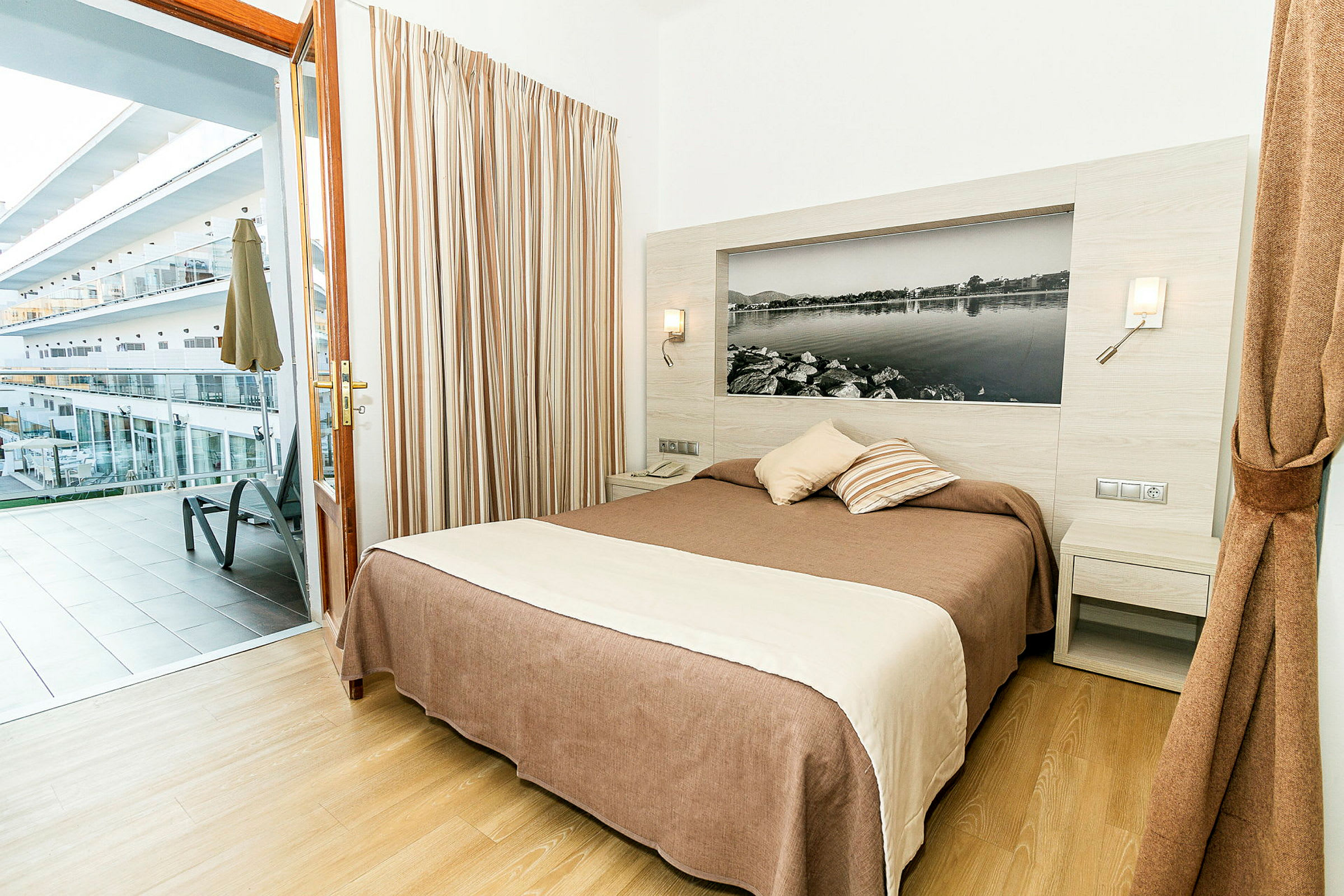 Ett hotellrum med en dubbelsäng och utgång till en balkong med utsikt över andra byggnader. (AI-genererad text)