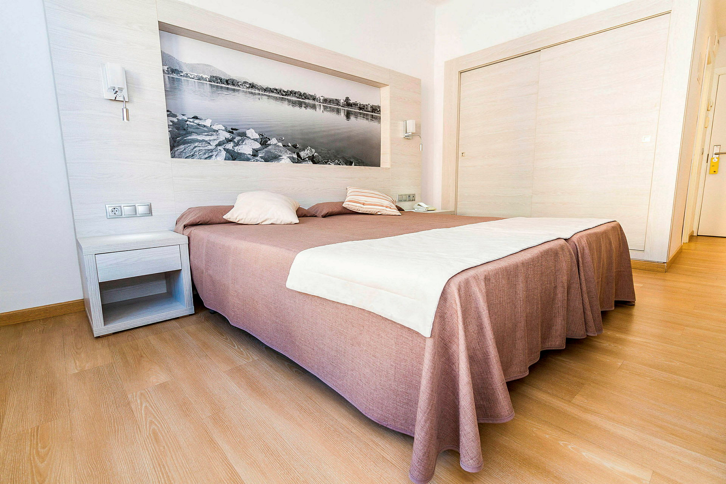 Hotellrum med en stor säng täckt av ett brunaktigt sängöverkast och vita lakan, med trägolv och en väggdekoration i form av en svartvit bild. (AI-genererad text)