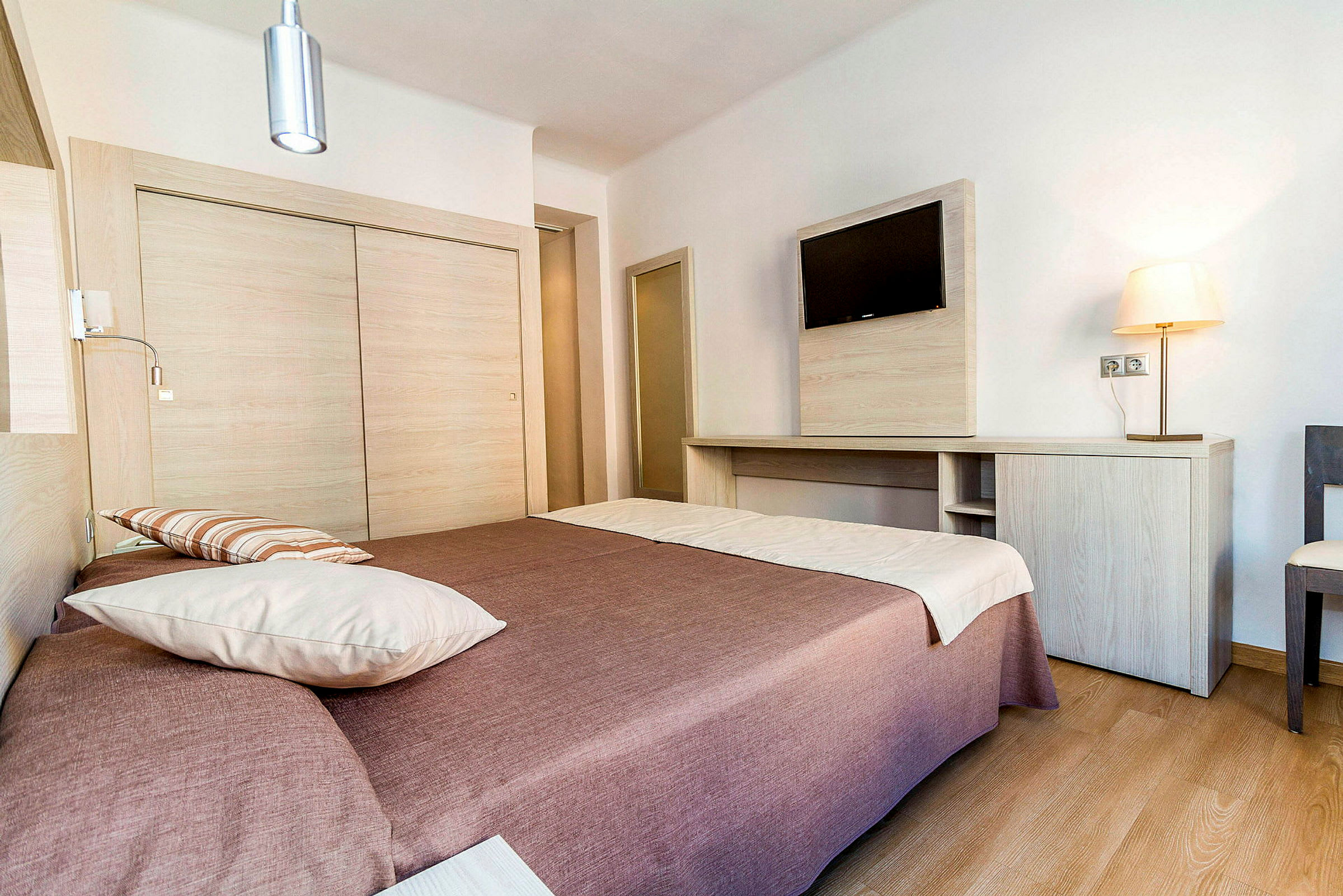 Ett hotellrum med en dubbelsäng täckt med brunt överkast och vita kuddar. På väggen hänger en platt-TV ovanför en träskrivbord. Rummet är ljust och modernt inrett. (AI-genererad text)