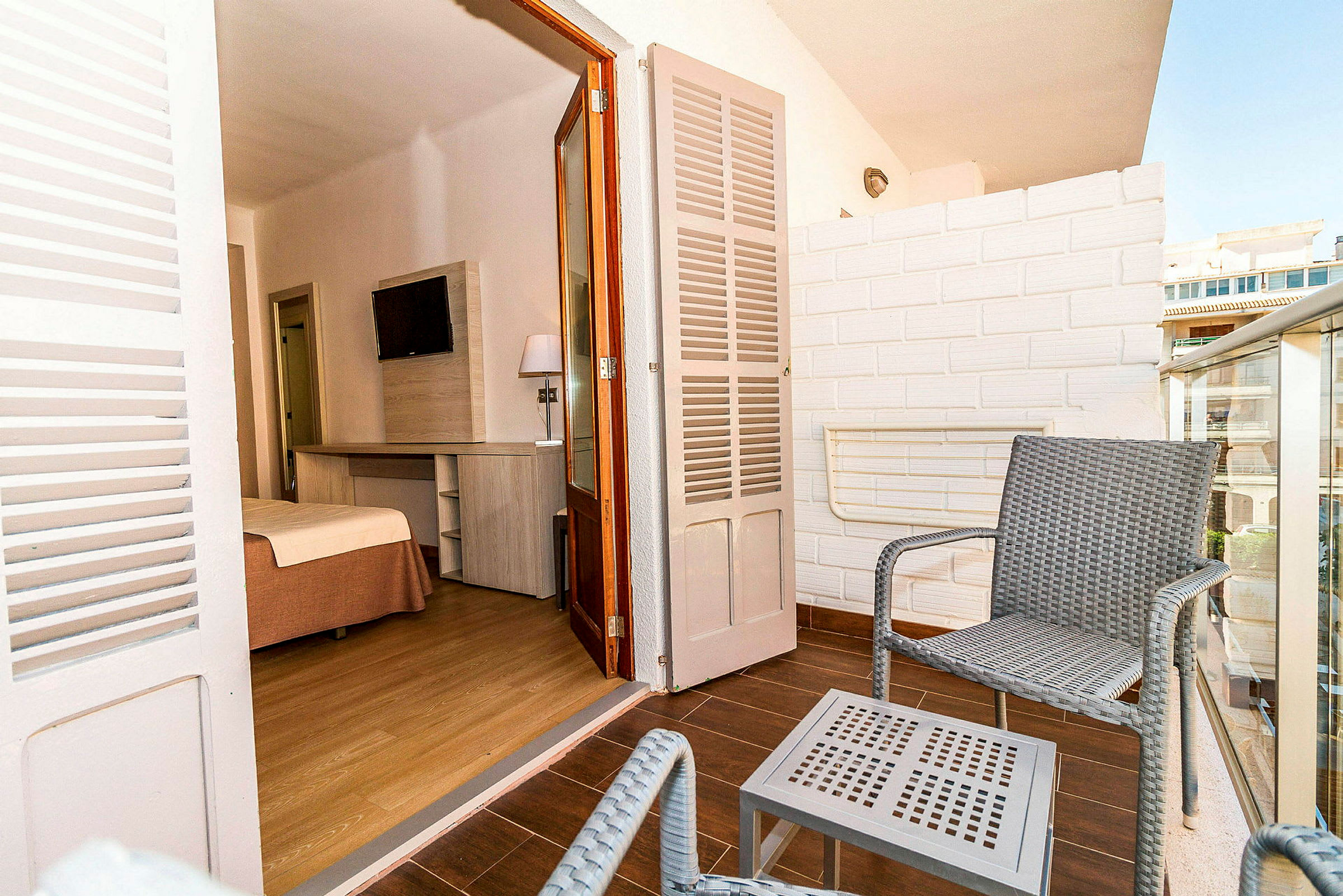 En balkong med två stolar och ett litet bord i flätad stil. I bakgrunden syns ingången till ett hotellrum med en säng och TV. (AI-genererad text)