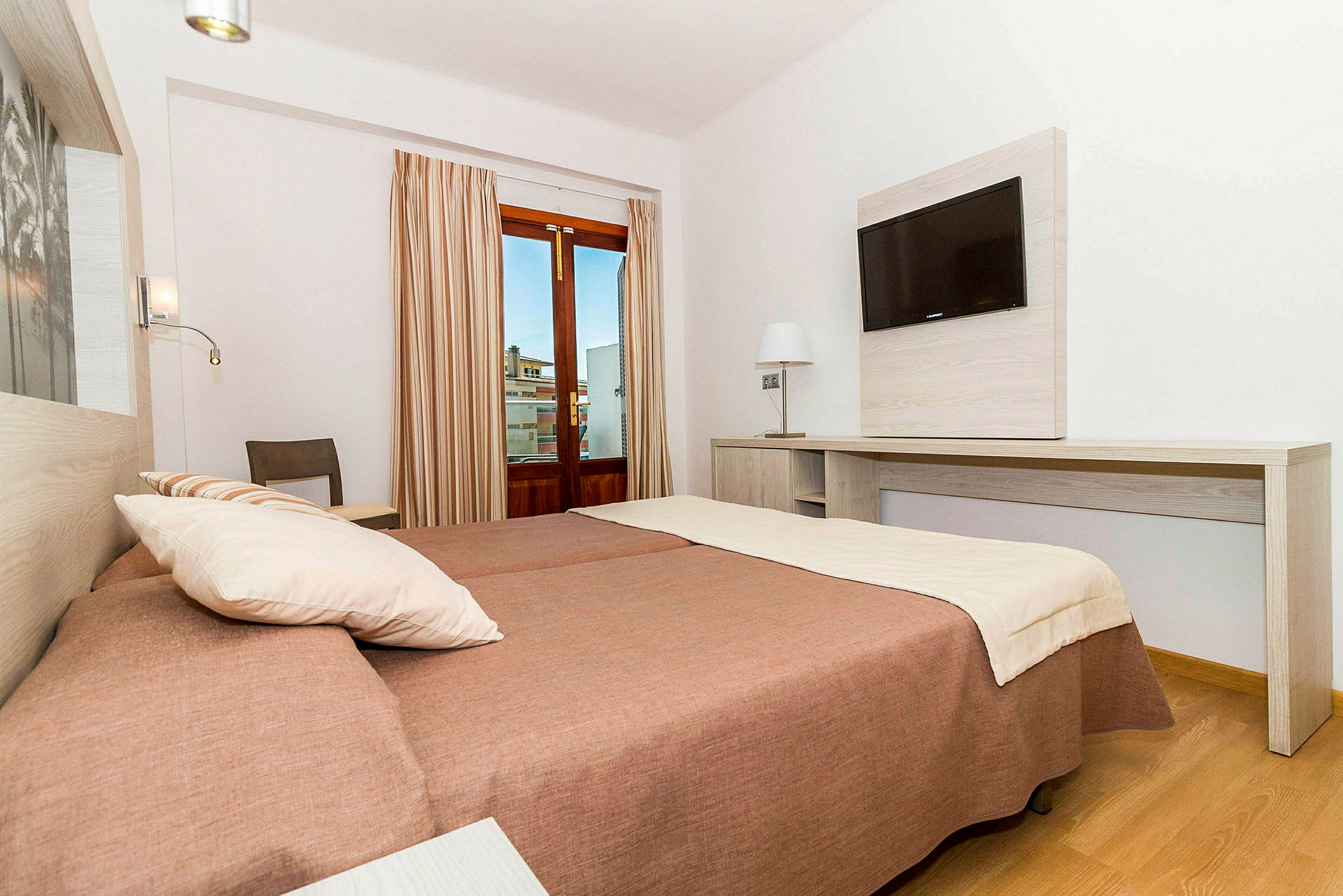 Ett hotellrum med två bäddar, skrivbord och väggmonterad TV samt ett fönster med beige gardiner. (AI-genererad text)