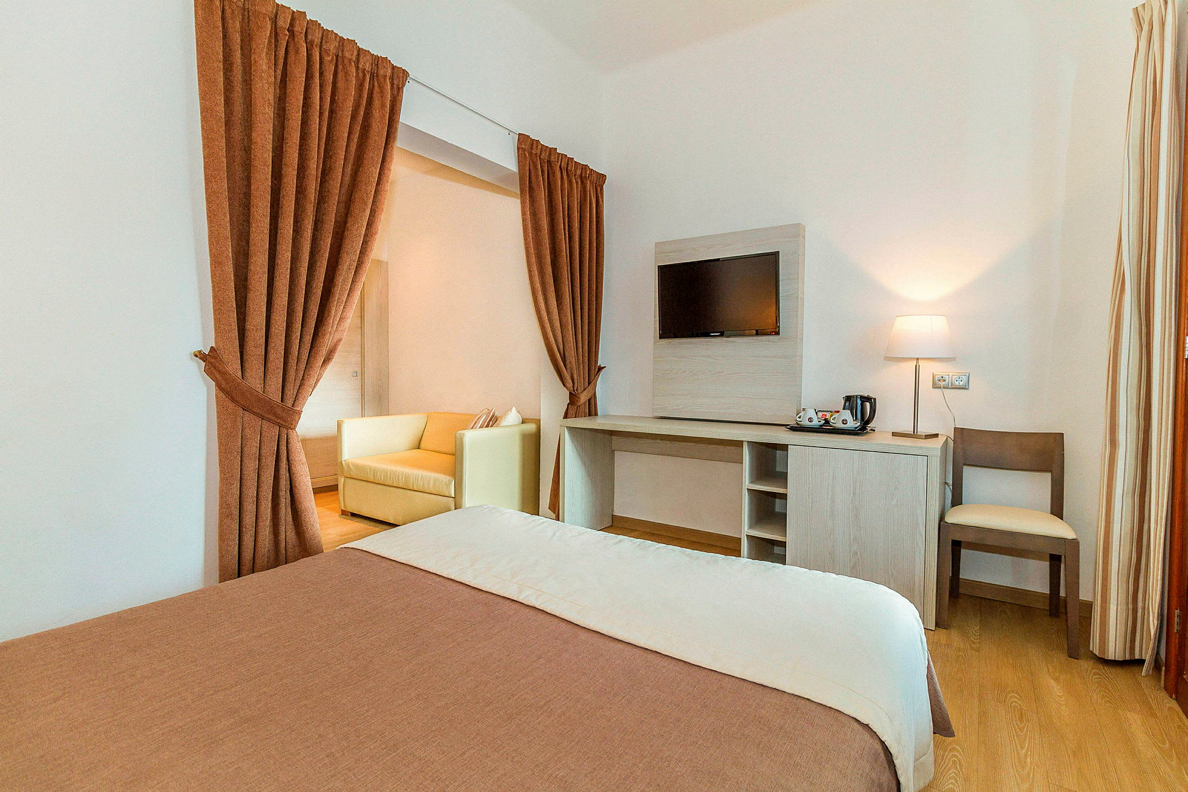 Ett hotellrum med en bäddad säng, en öppen sittgrupp bakom ett draperi och ett skrivbord med en stol intill en vägg. (AI-genererad text)