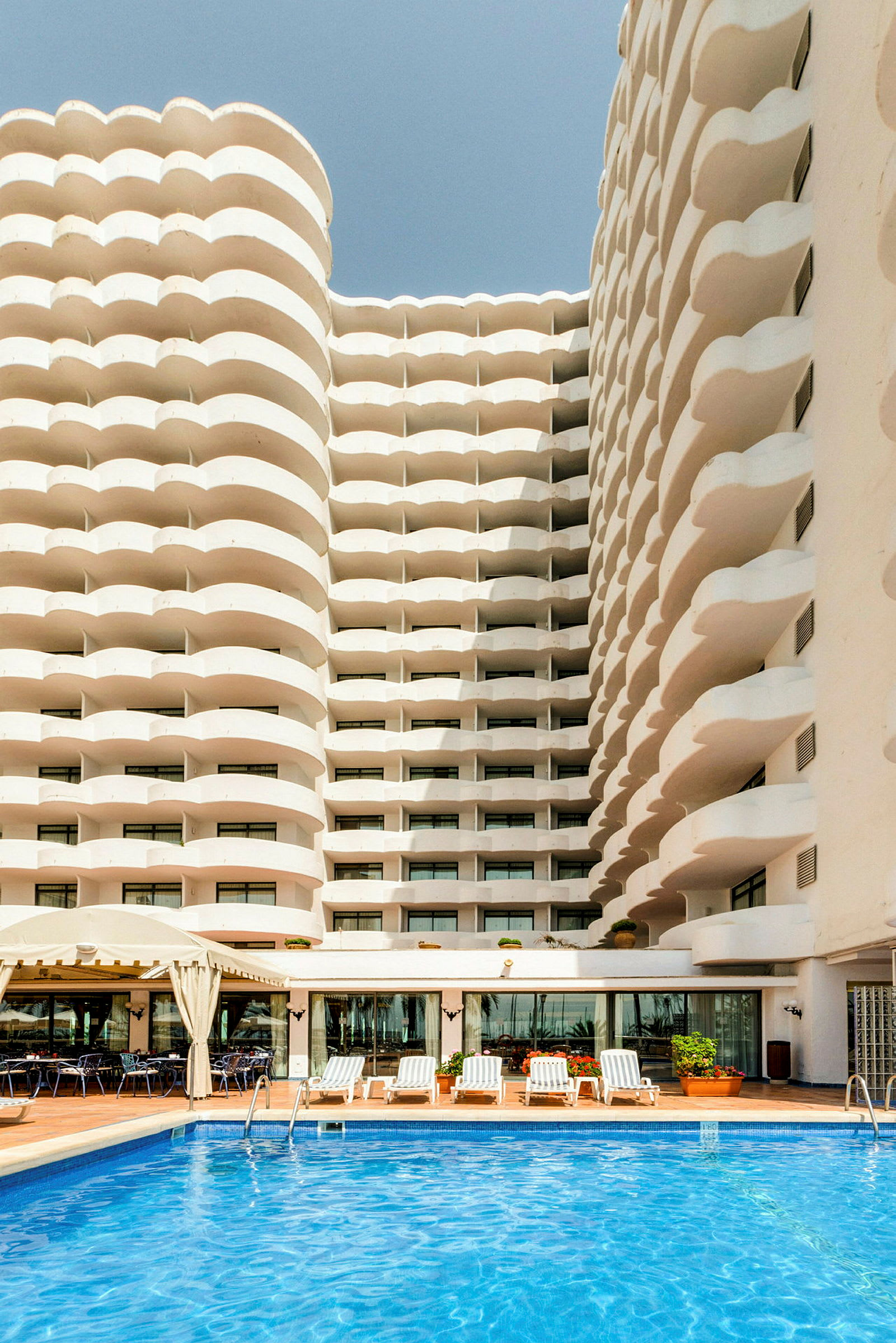 Framsidan av ett hotell med vågformade balkonger och en pool i förgrunden. (AI-genererad text)