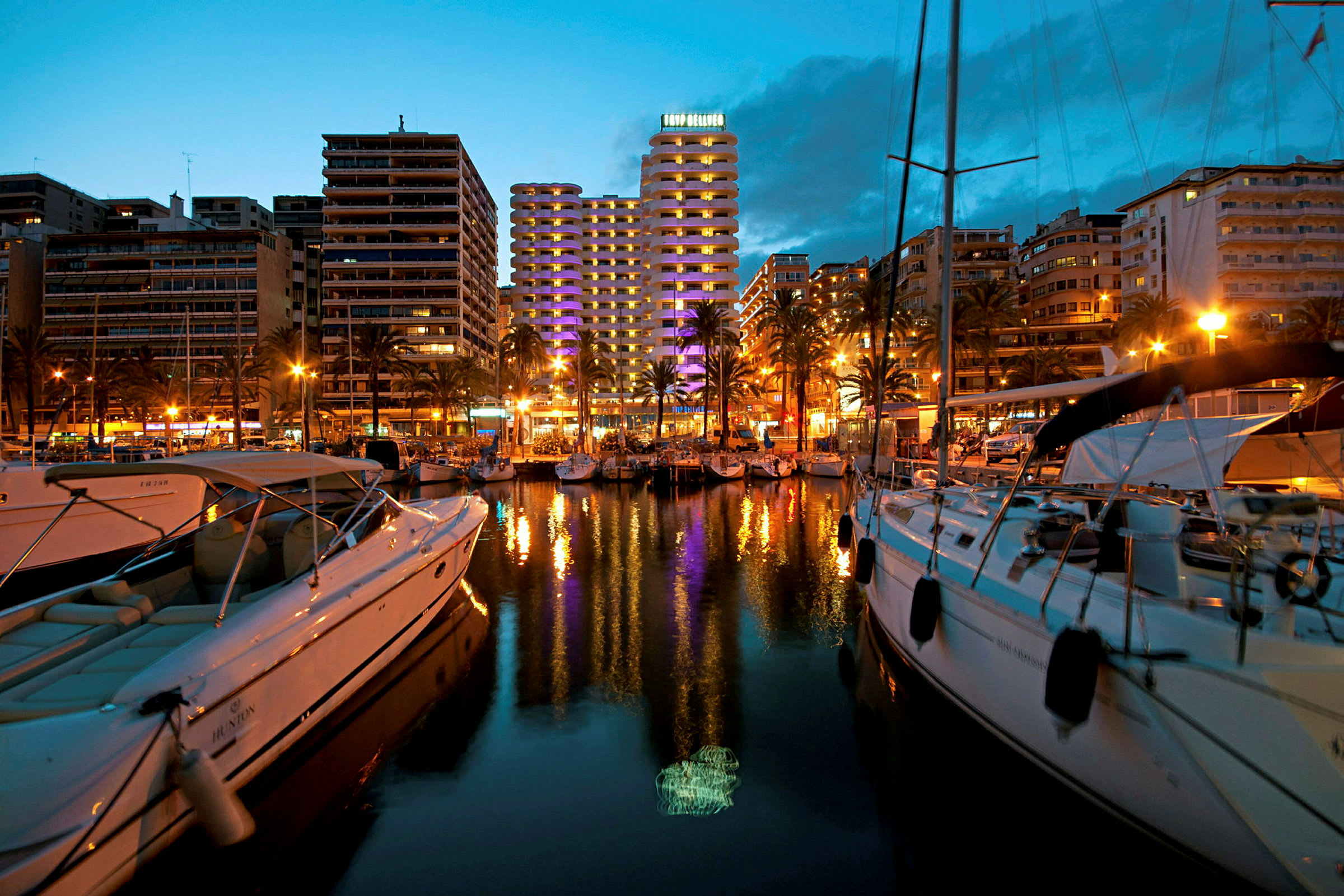 En marina i en stad, omgiven av stora byggnader med belysning, båtar och vatten som speglar ljusen. (AI-genererad text)
