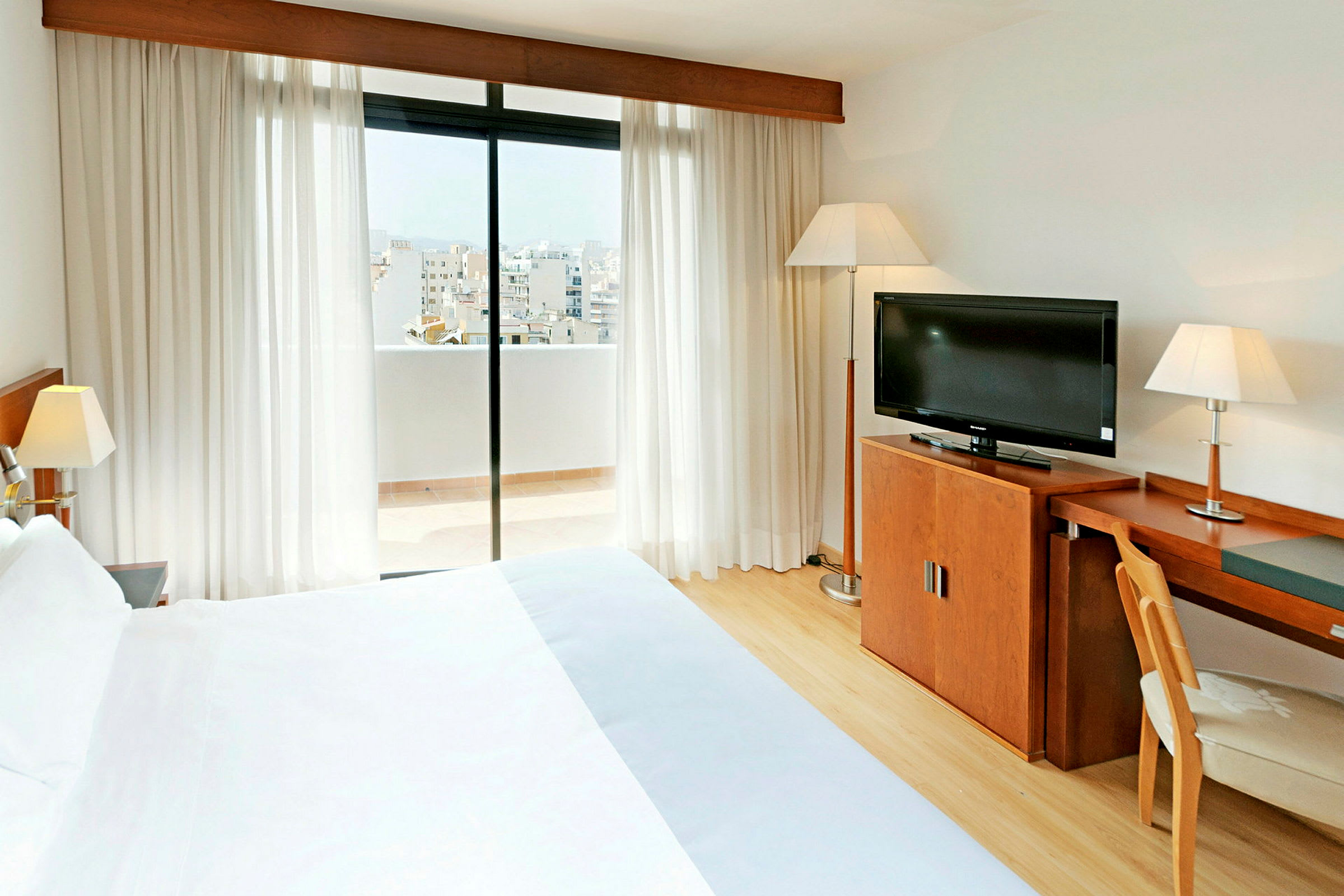 Ett hotellrum med en dubbelsäng, skrivbord, TV och en balkong med utsikt över staden. (AI-genererad text)
