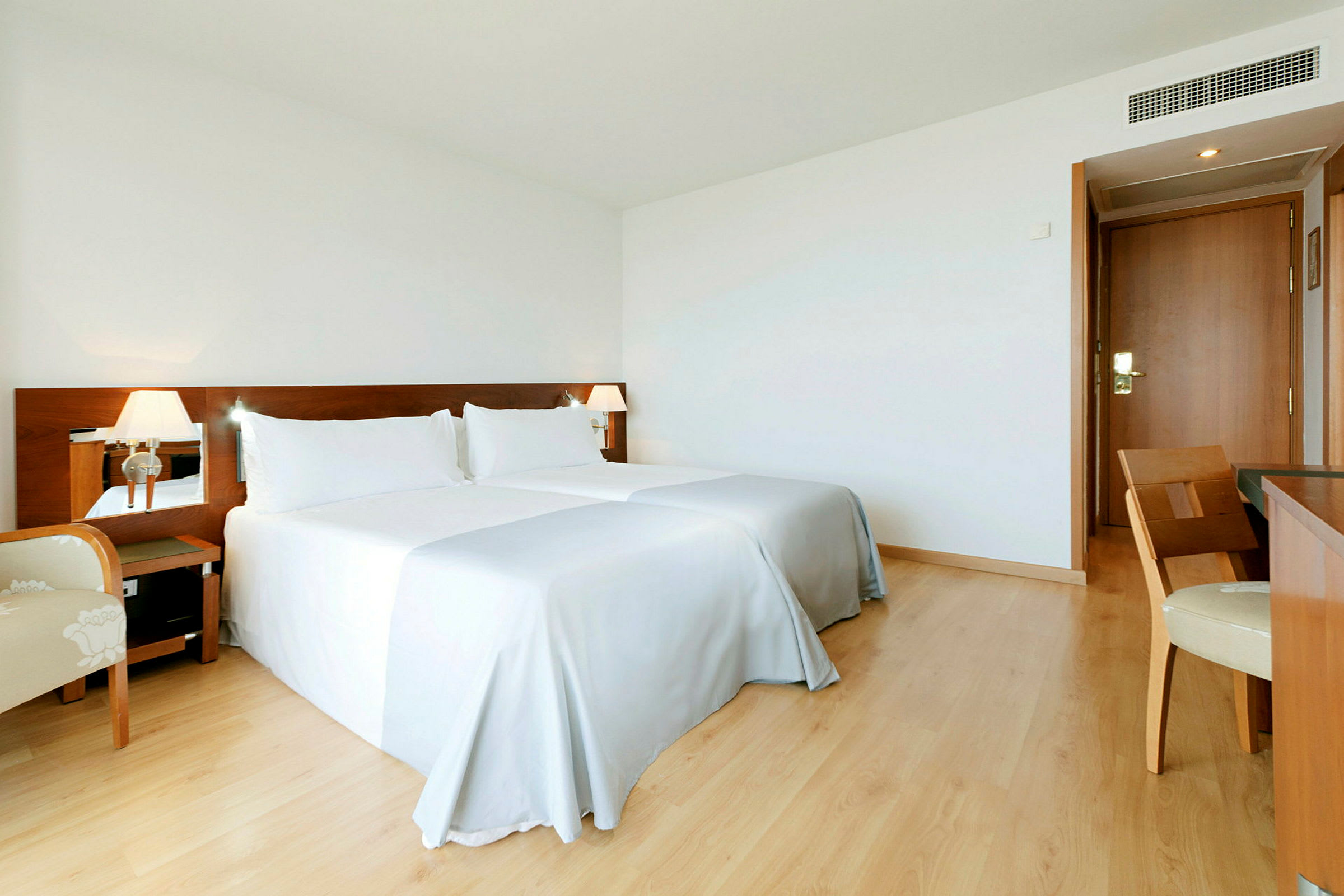 Ett hotellrum med två sängar, vita sängkläder och en träpanel bakom sängarna. Det finns ett skrivbord med en stol och en dörr i bakgrunden. (AI-genererad text)