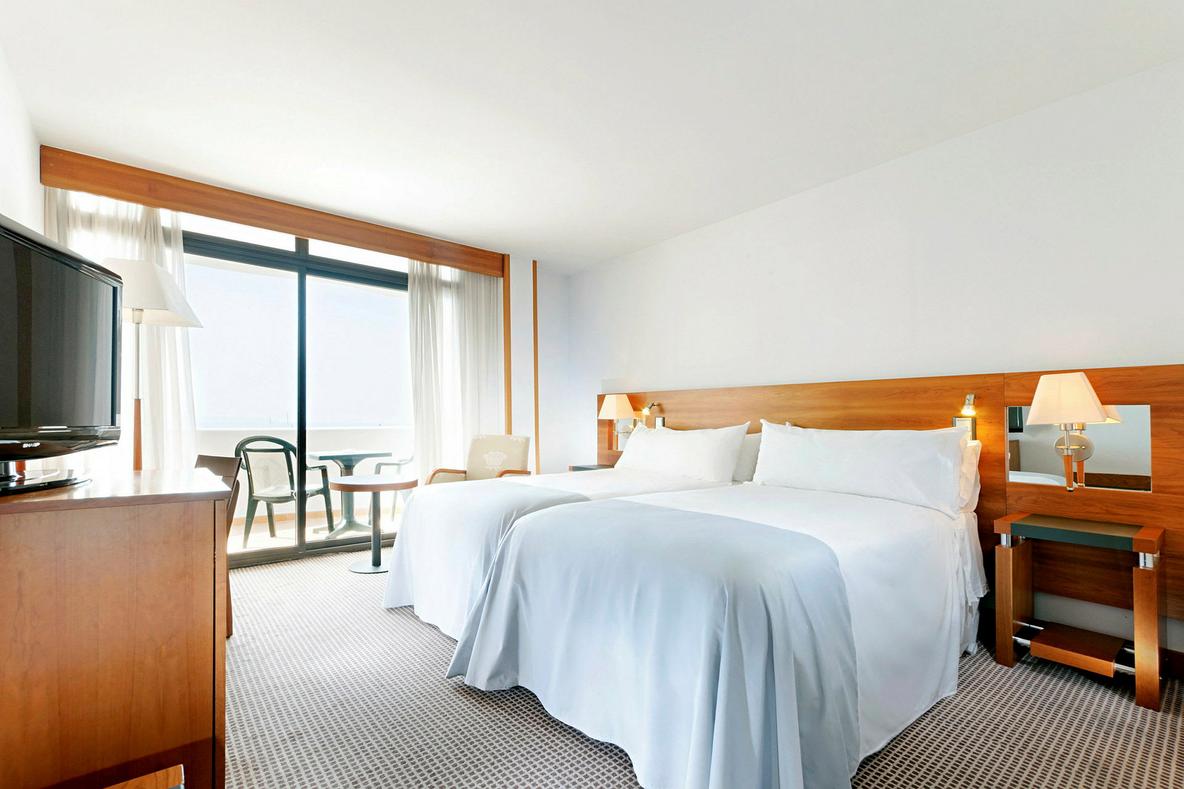 Ett ljus, rymligt hotellrum med en dubbelsäng, trämöbler, en balkong och utsikt genom stora fönster. (AI-genererad text)