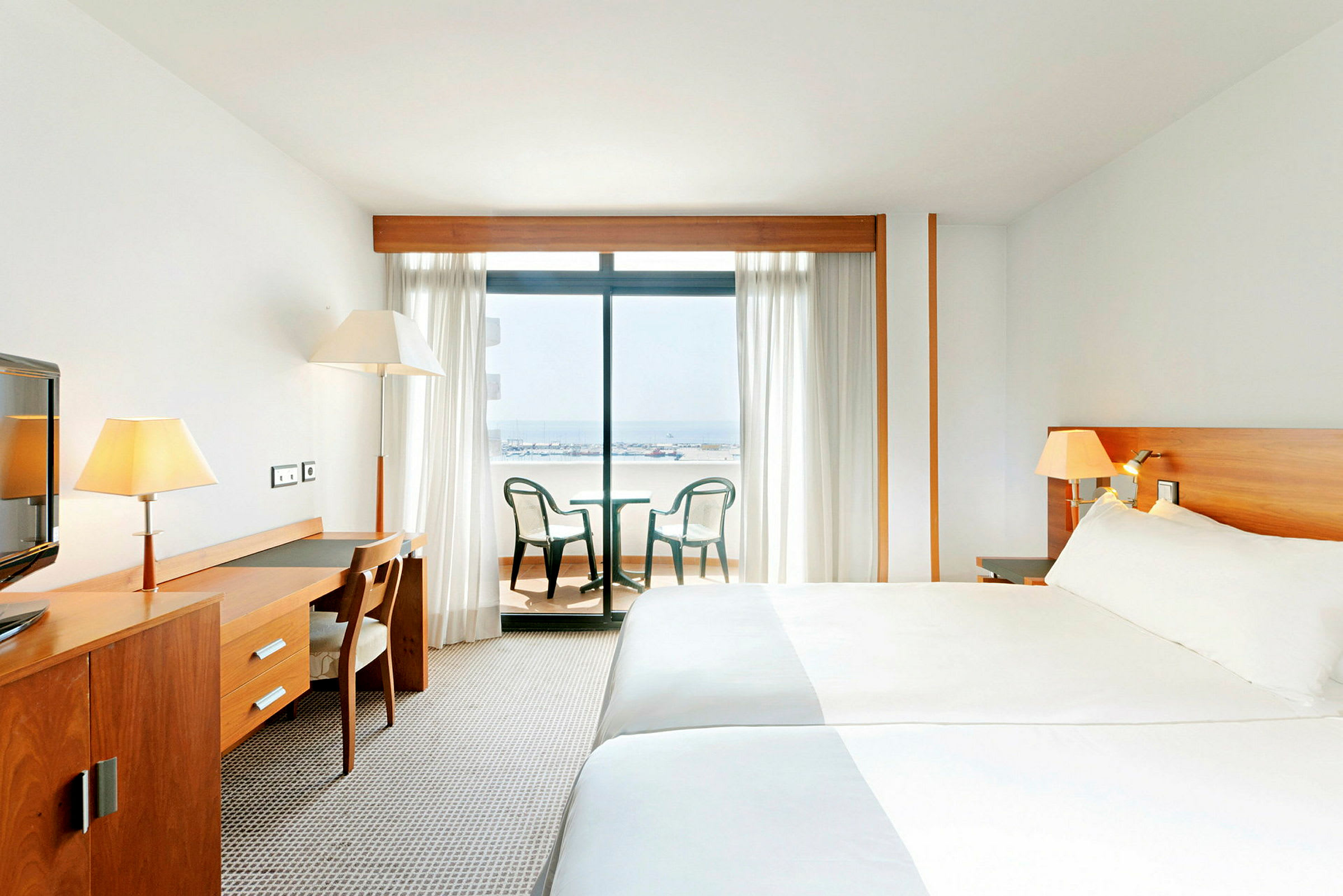 Hotellrum med en stor säng, ett skrivbord med stol, och balkongmöbler framför en glasdörr som visar en utsikt över havet. (AI-genererad text)