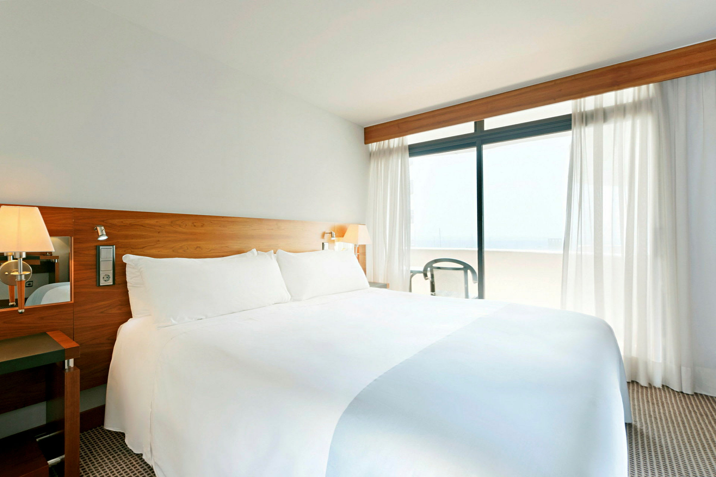 Ett ljust hotellrum med en stor bäddad säng och vitt sänglinne, ett trähuvudgavel och fönster med gardiner som leder till en balkong. (AI-genererad text)