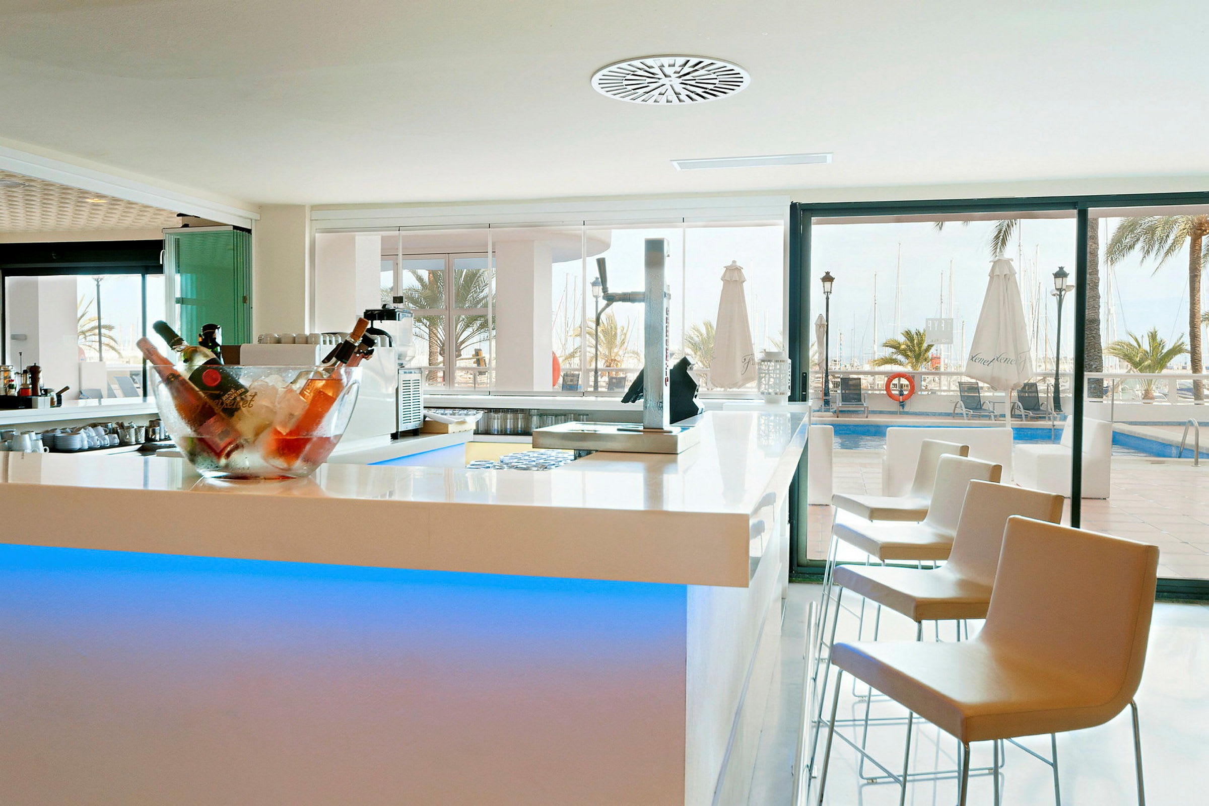Bild av en modern bar med ljusa möbler, en skål fylld med flaskor på is och utsikt över en pool och marina i bakgrunden. (AI-genererad text)