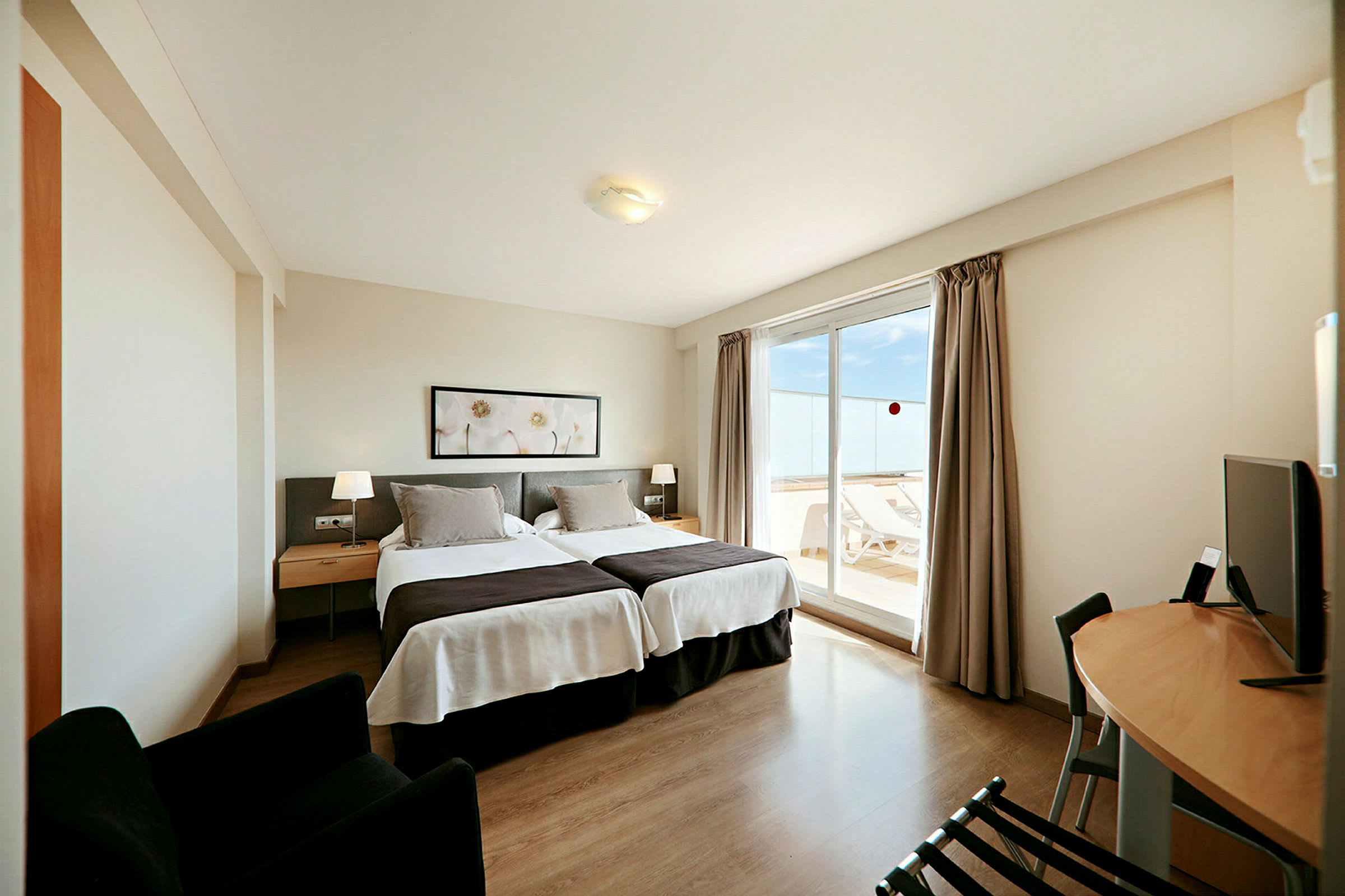 Ett hotellrum med två sängar, en liten sittgrupp och ett skrivbord. Rummet har stora fönster som leder till en balkong med solstolar. (AI-genererad text)