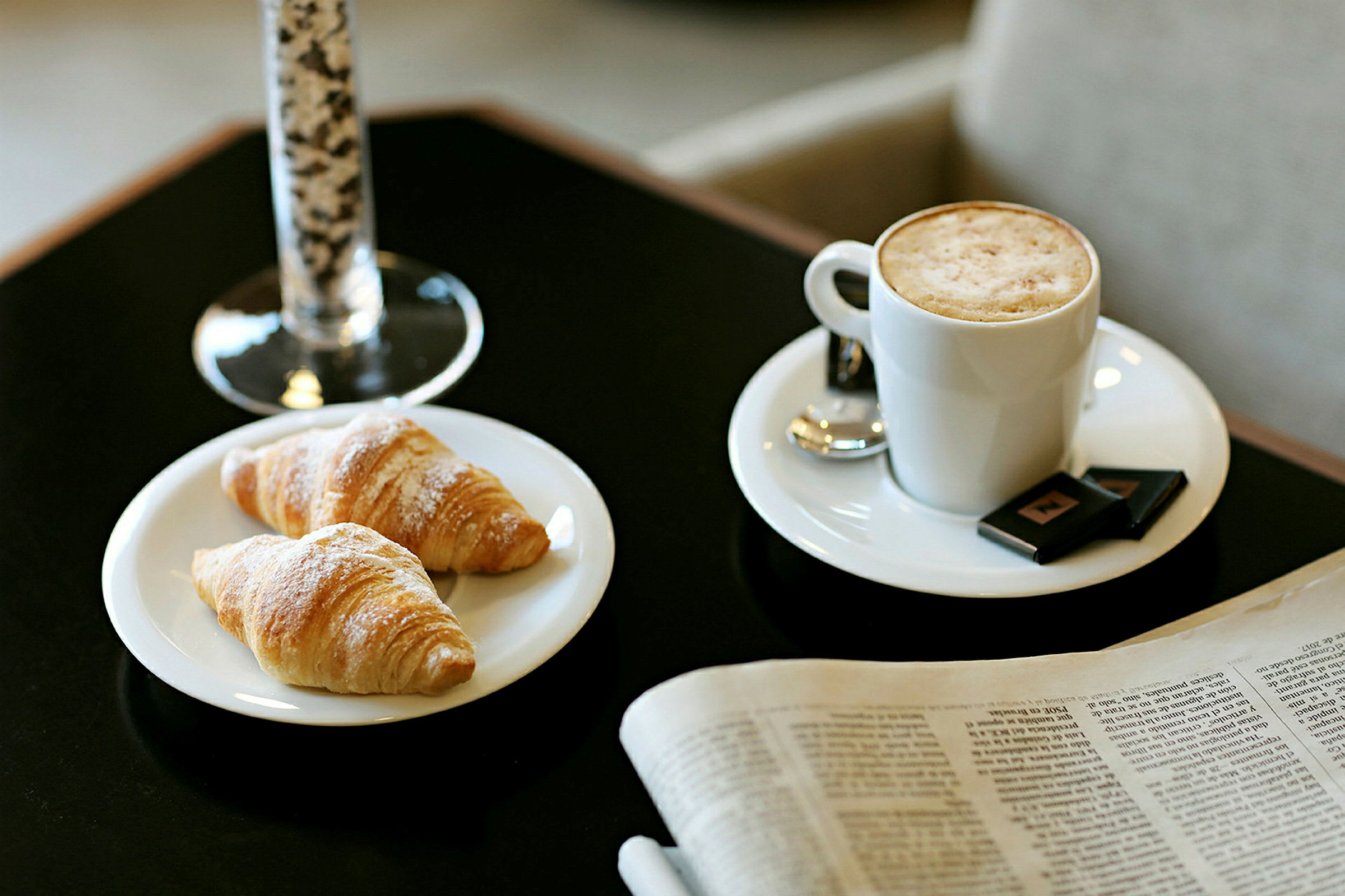 Bild föreställer två croissanter på en tallrik, en kopp kaffe med mjölkskum och ett glas i bakgrunden. En tidning ligger bredvid på bordet. (AI-genererad text)