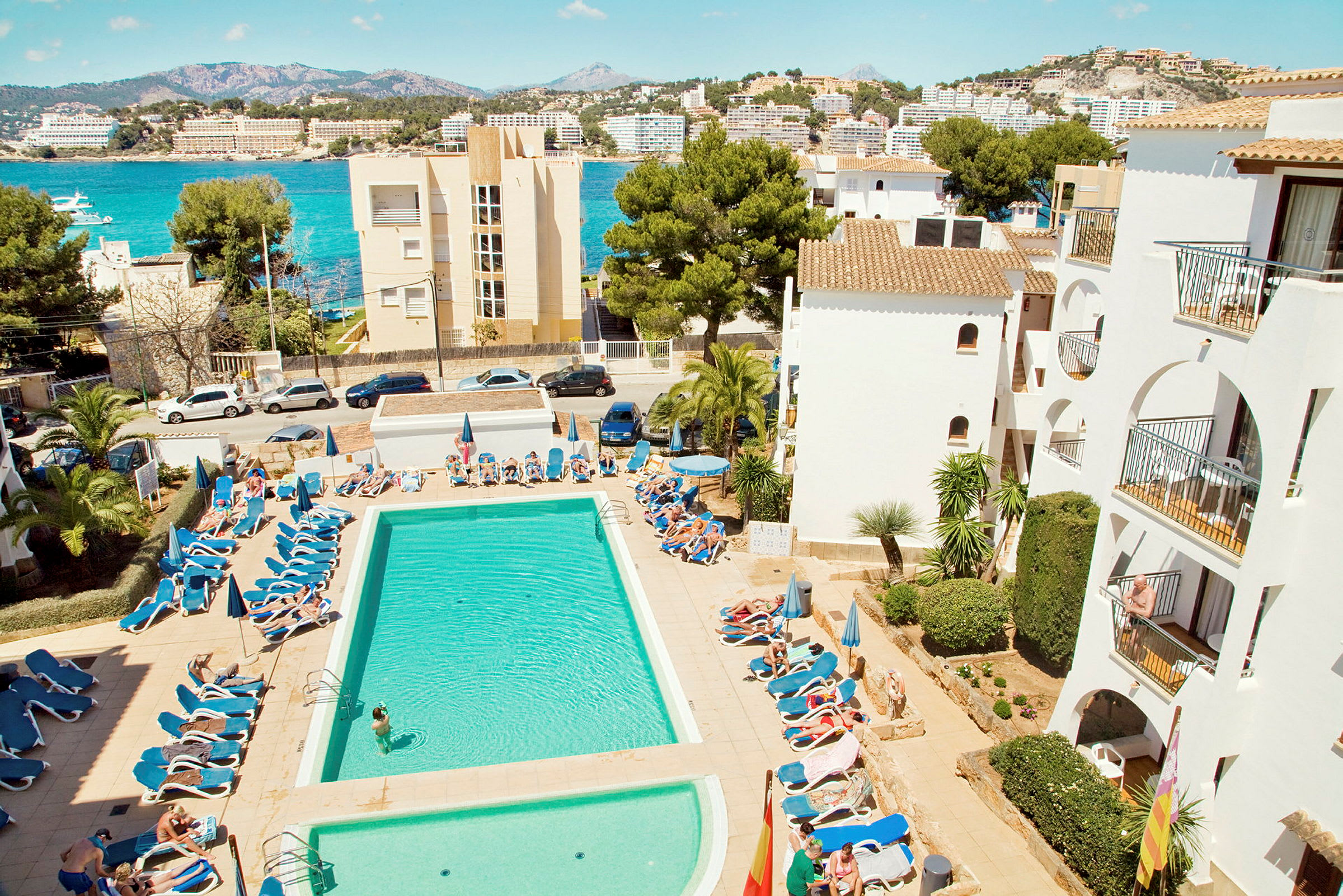 En pool med solstolar omgiven av vita byggnader, med utsikt över hav och kullar. (AI-genererad text)