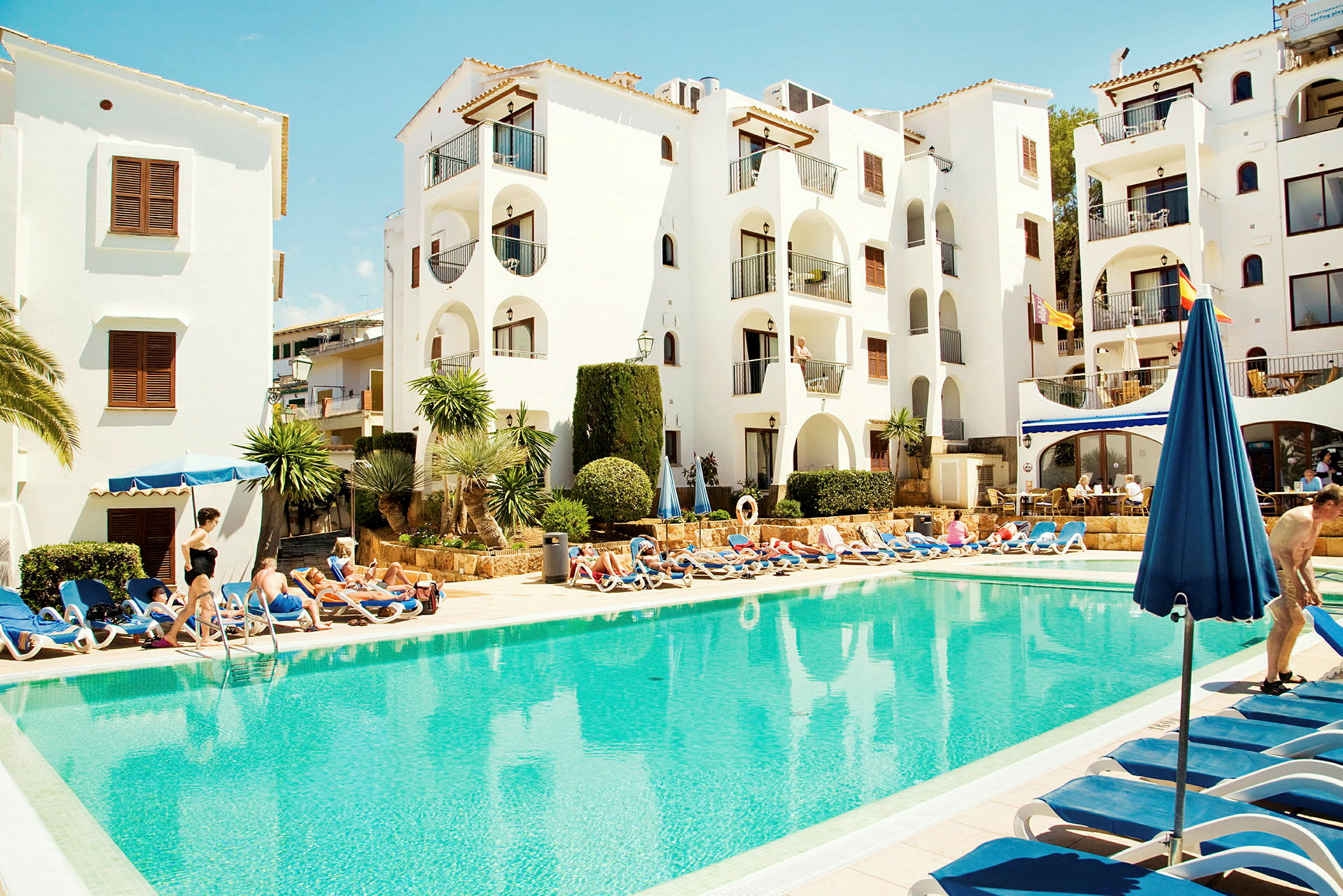 Ett poolområde med solstolar runt en klarblå pool, framför en vit byggnad med balkonger. (AI-genererad text)