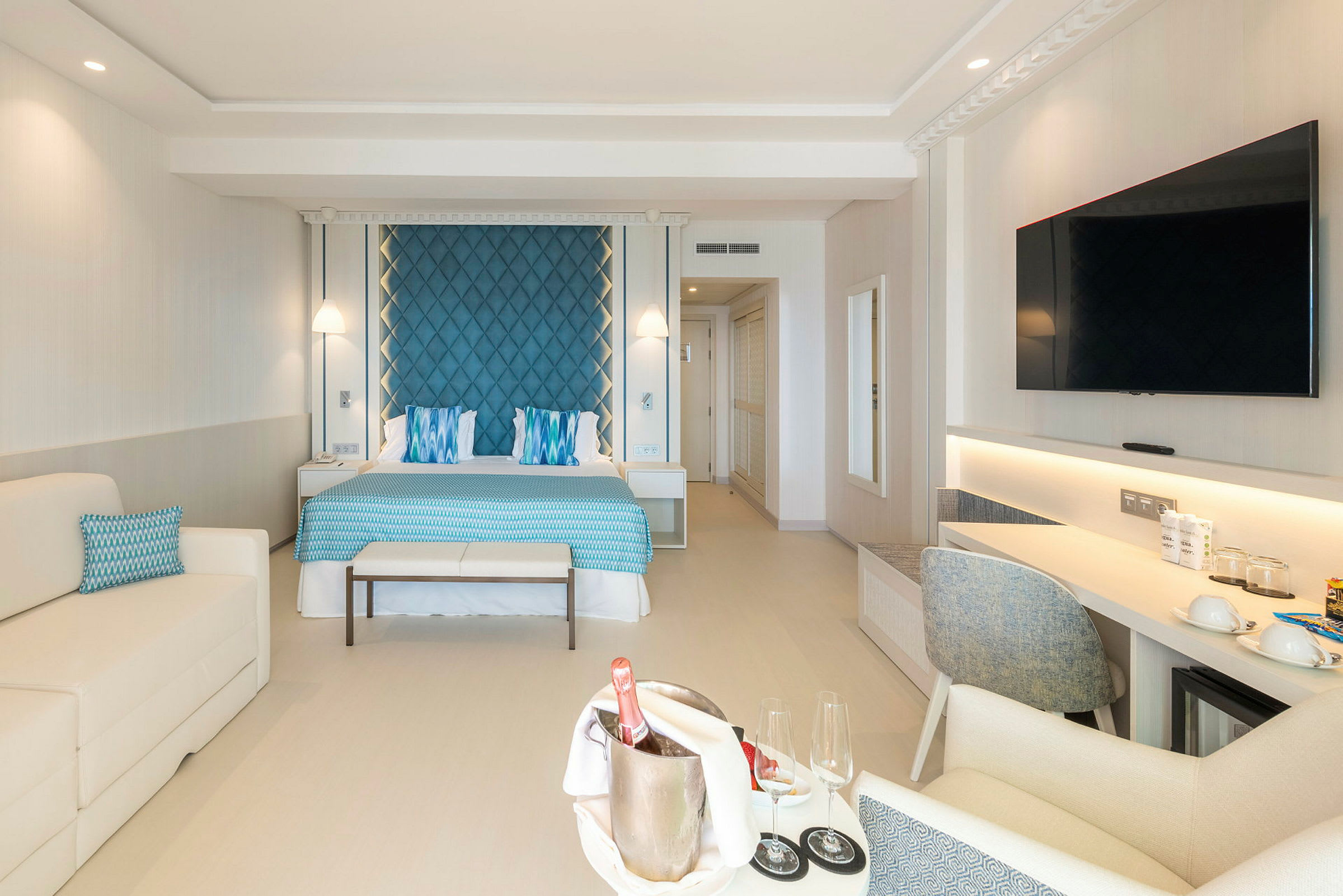 Ett hotellrum med en dubbelsäng, blå sänggavel och vita möbler, samt ett bord med champagne och glas. (AI-genererad text)