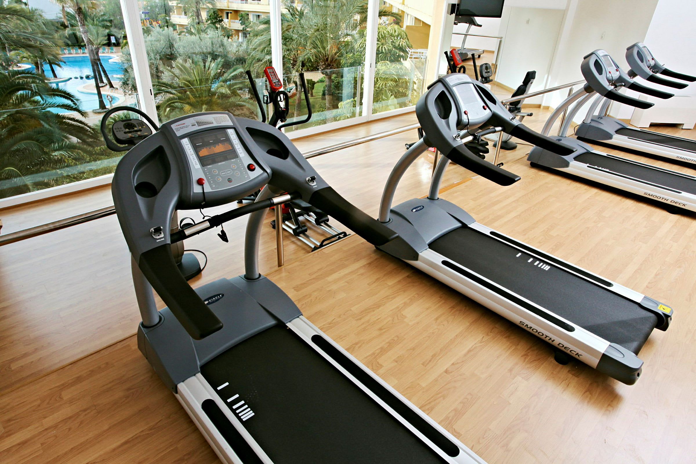 Bild på ett gym med löpband och träningsutrustning framför stora fönster med utsikt över en pool och palmer. (AI-genererad text)