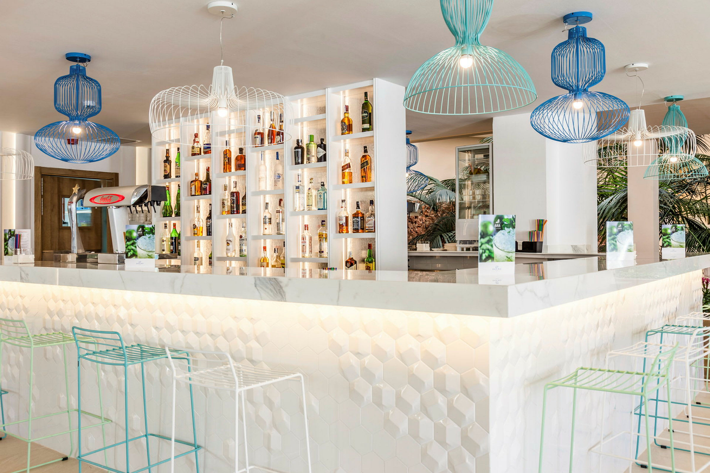 En modern bar med vita hyllor fyllda med flaskor, geometriska lampor i blått och grönt hänger i taket, samt stolar med smala metallramar vid bardisken. (AI-genererad text)