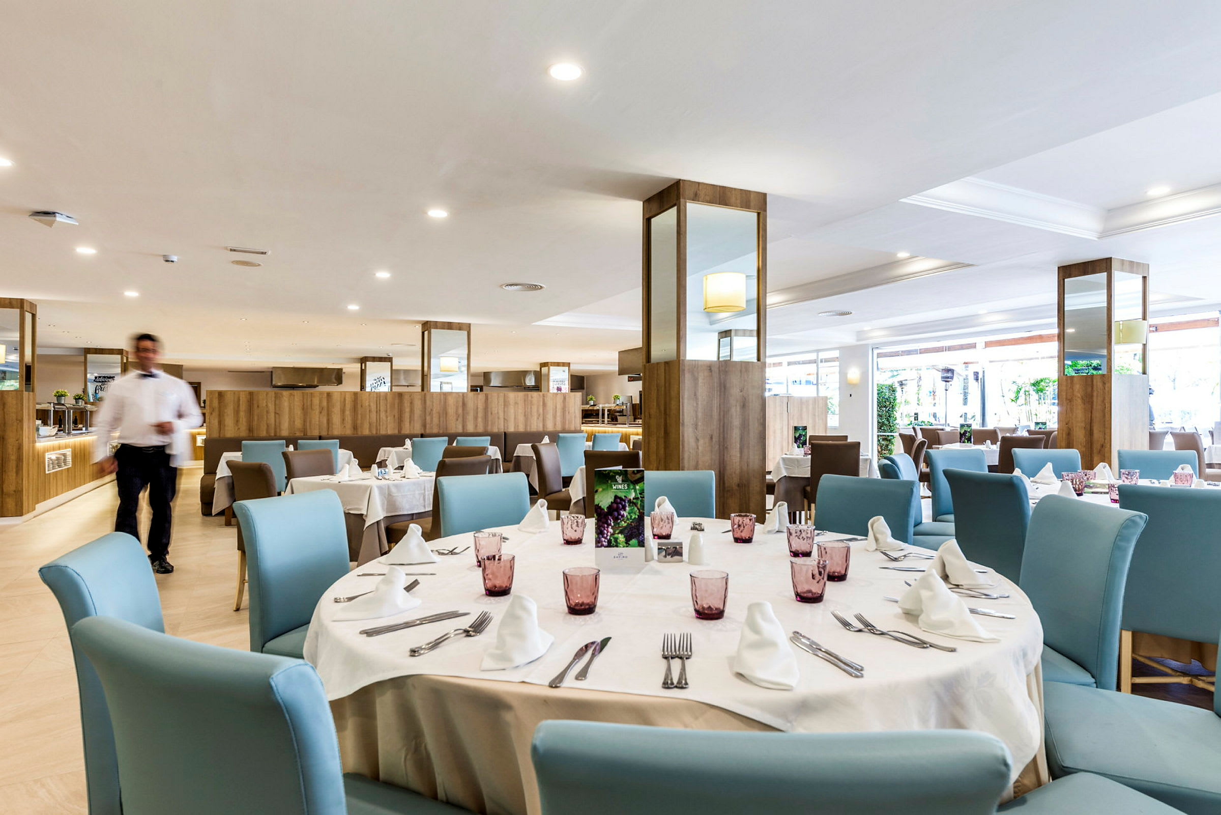 En restaurang med dukade bord, ljusa blå stolar och en servitör som rör sig i rummet. (AI-genererad text)