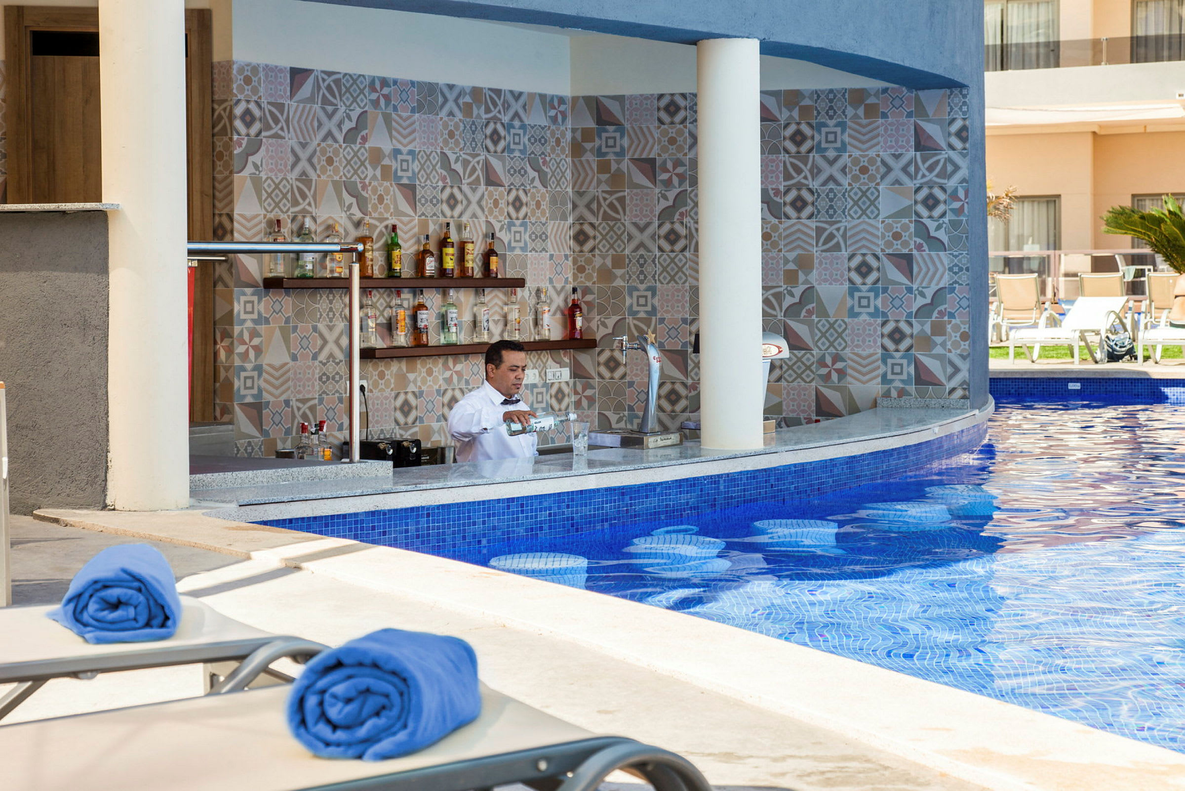 En bild på en poolbar med en bartender som gör en drink. Blåa handdukar ligger på solstolar vid poolområdet. (AI-genererad text)