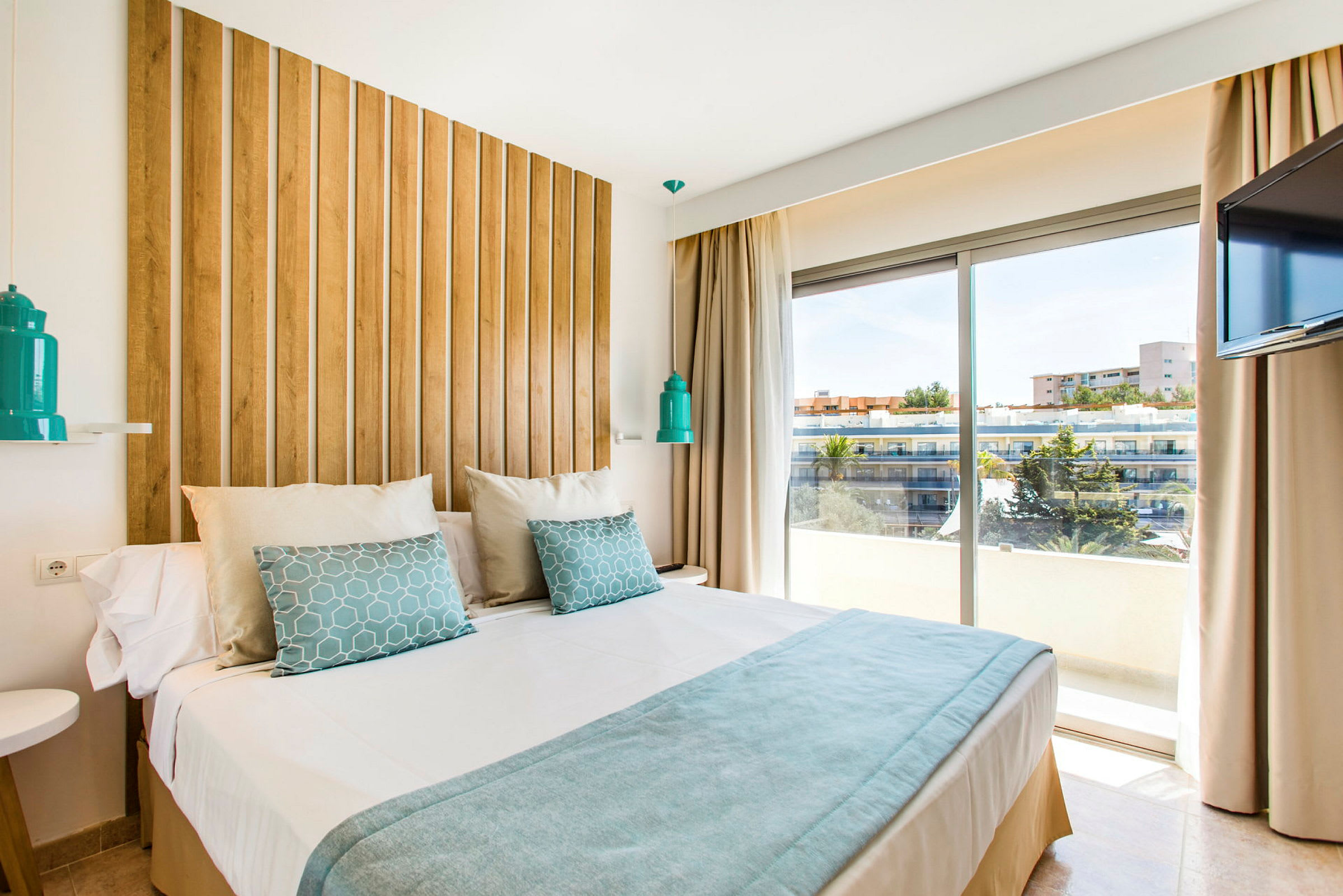 Ett modernt hotellrum med en stor säng, träpanel bakom sängen, vita och blå sängkläder, samt en fönstervy över byggnader och träd utanför. (AI-genererad text)