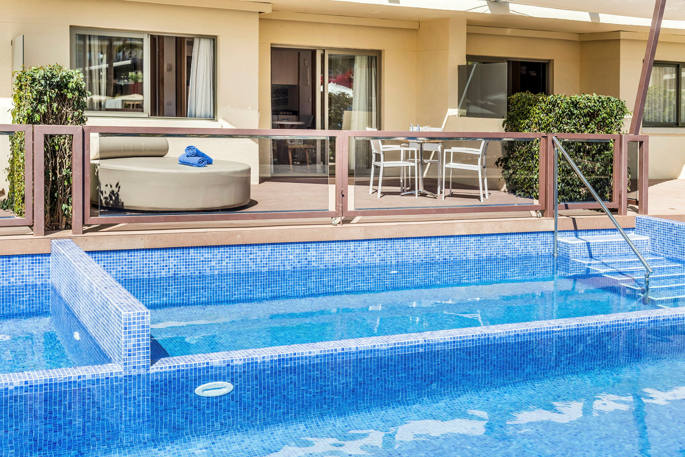 En pool med blå kakel framför en uteplats med möbler, inklusive en sittplats med blåa handdukar och ett bord med stolar. (AI-genererad text)