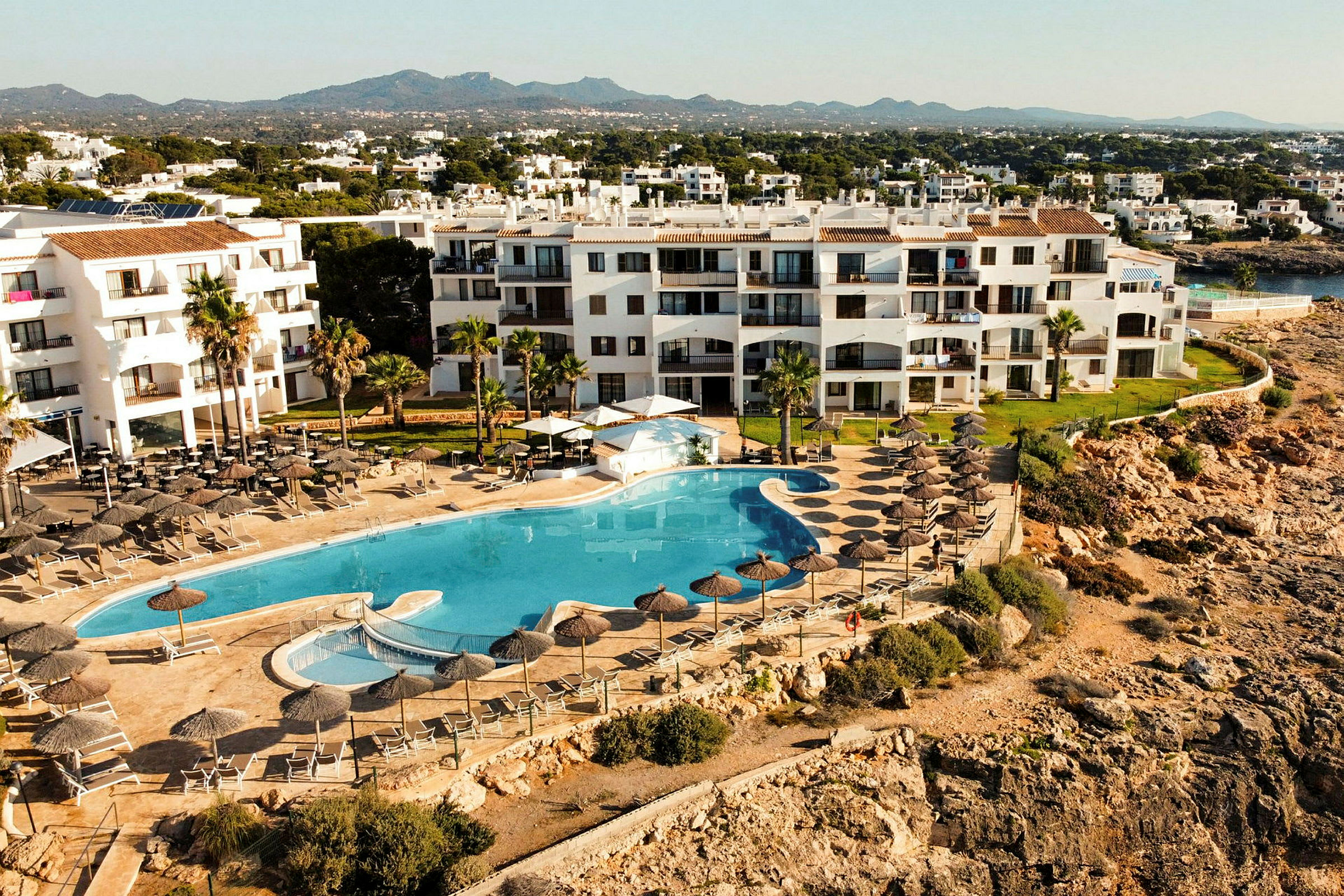 Hotellbyggnad vid klippor med stor pool och solstolar. (AI-genererad text)