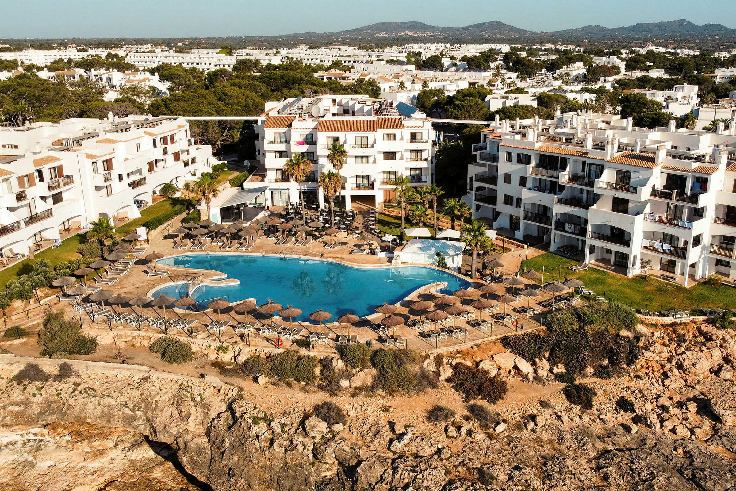 Hotell med vit fasad vid klippkant med pool och solstolar. (AI-genererad text)
