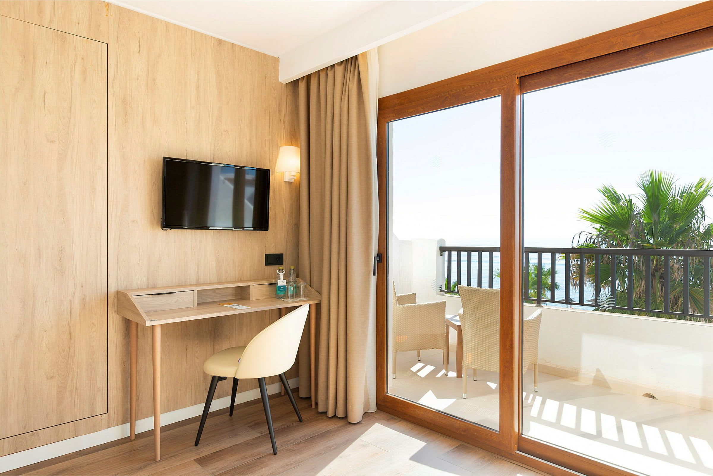 Hotellrum med träpanel, skrivbord, stol och en TV på väggen. Stora fönster med utsikt mot en balkong med möbler och palmer. (AI-genererad text)