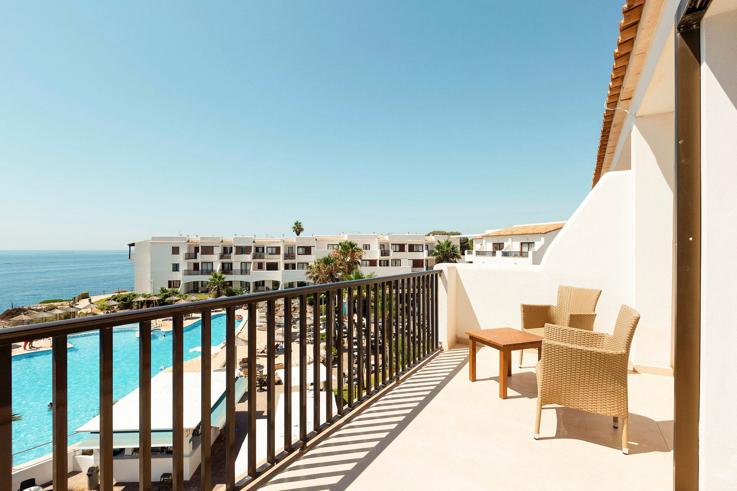 En balkong med två flätade stolar och ett bord, utsikt över ett hotellområde med utomhuspool och havet i bakgrunden. (AI-genererad text)