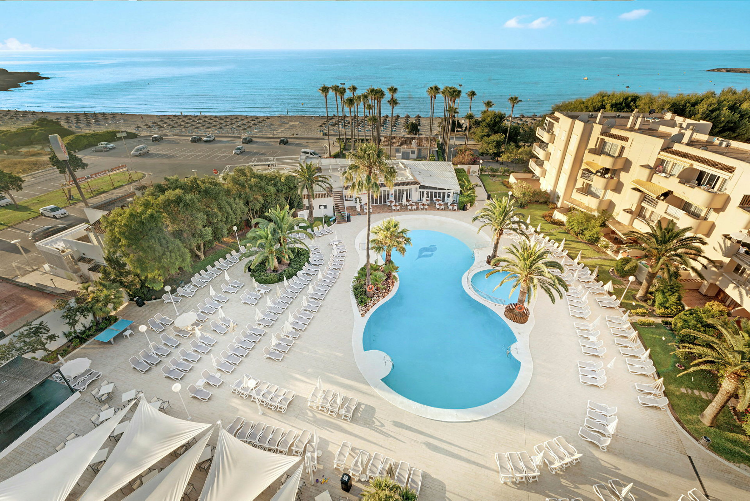 Flygbild över hotellområde med pool, solstolar och palmer, samt närhet till stranden. (AI-genererad text)