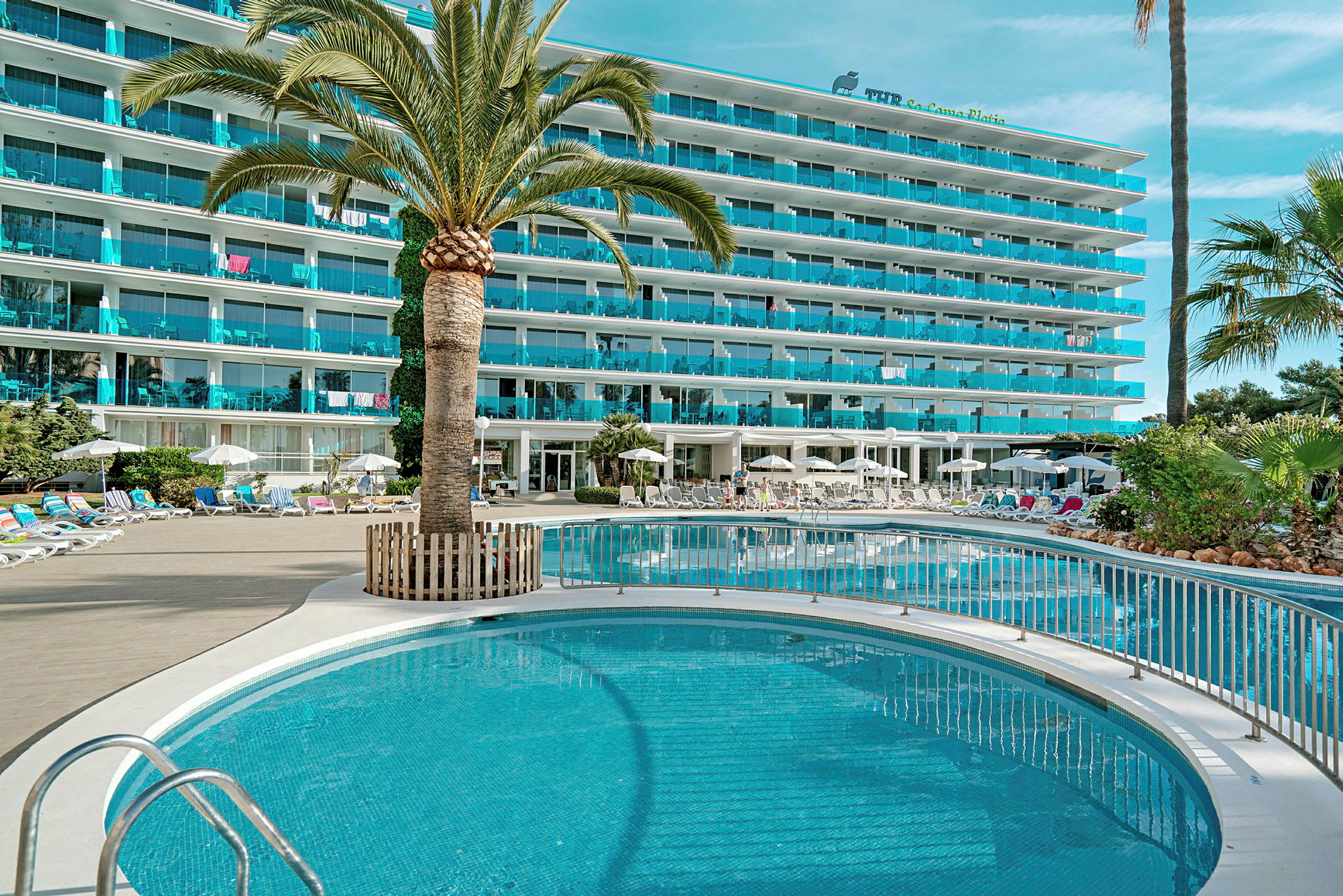 Bild av ett hotell med en pool, omgiven av solstolar och parasoller, med palmer och gröna växter. (AI-genererad text)