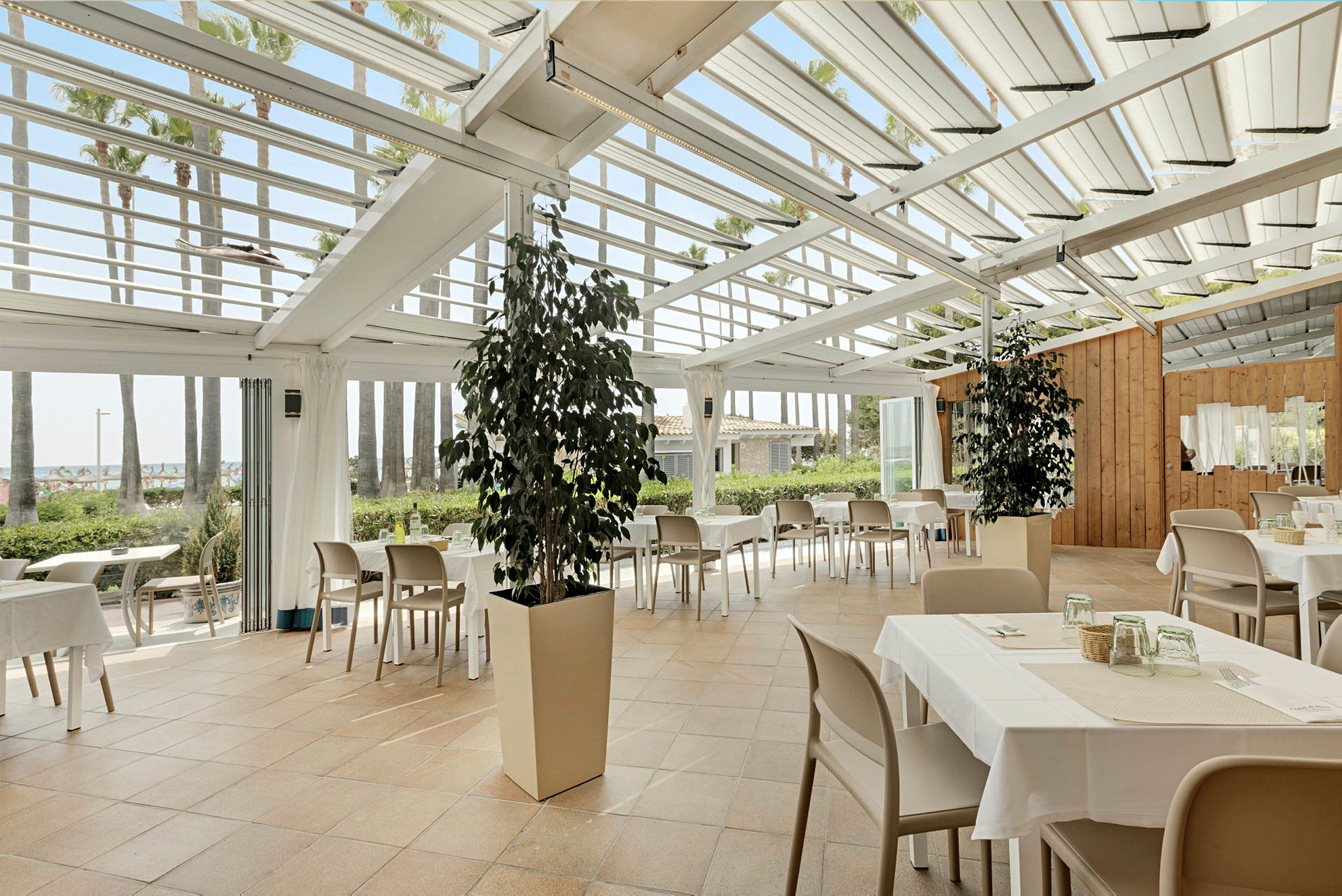 Restaurang med bord och stolar under ett glastak, omgiven av gröna växter och palmer. (AI-genererad text)