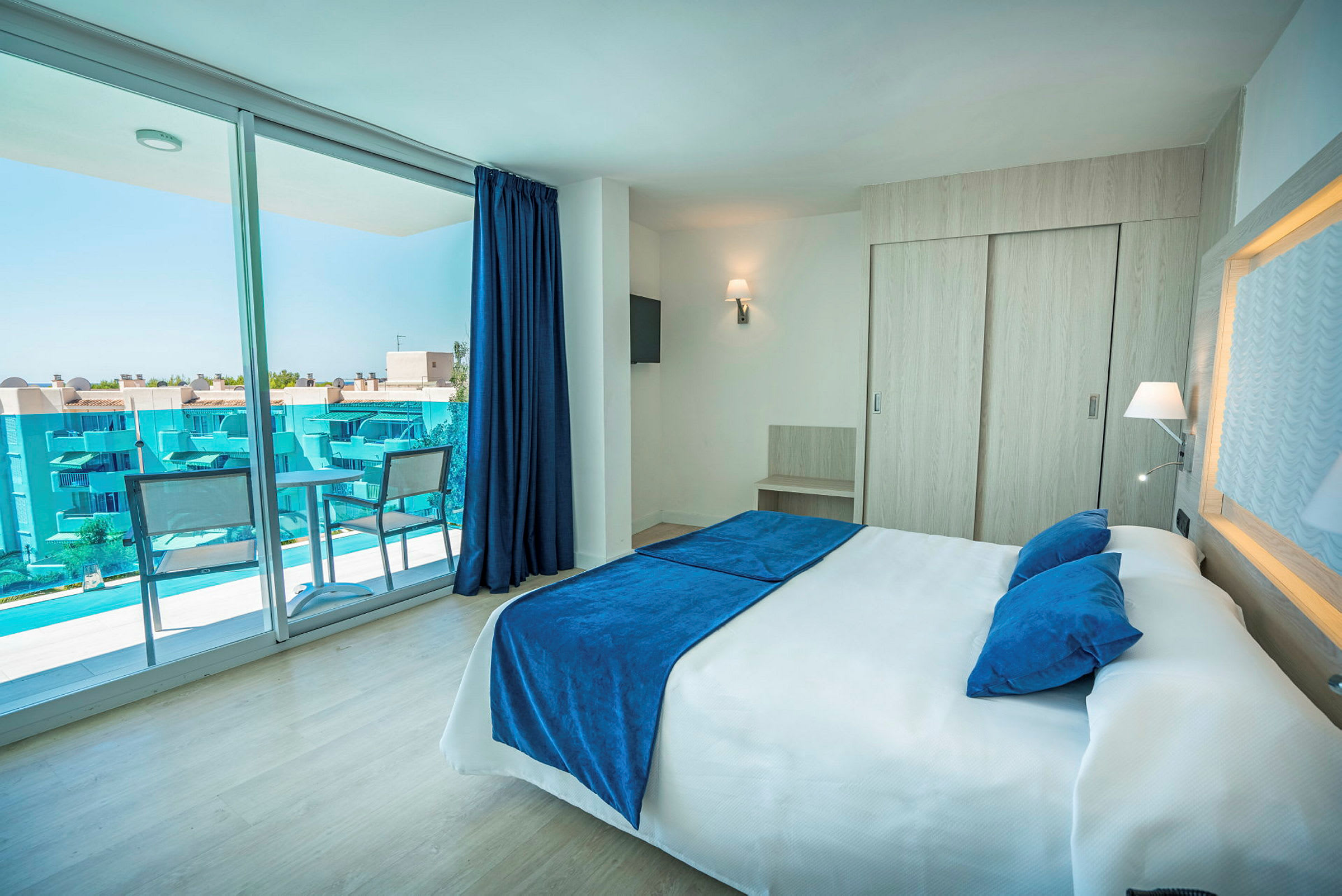 Ett hotellrum med en stor säng, blått överkast och kuddar. Rummet har även ett stort glasfönster som vetter mot en balkong med utsikt över blå byggnader. (AI-genererad text)