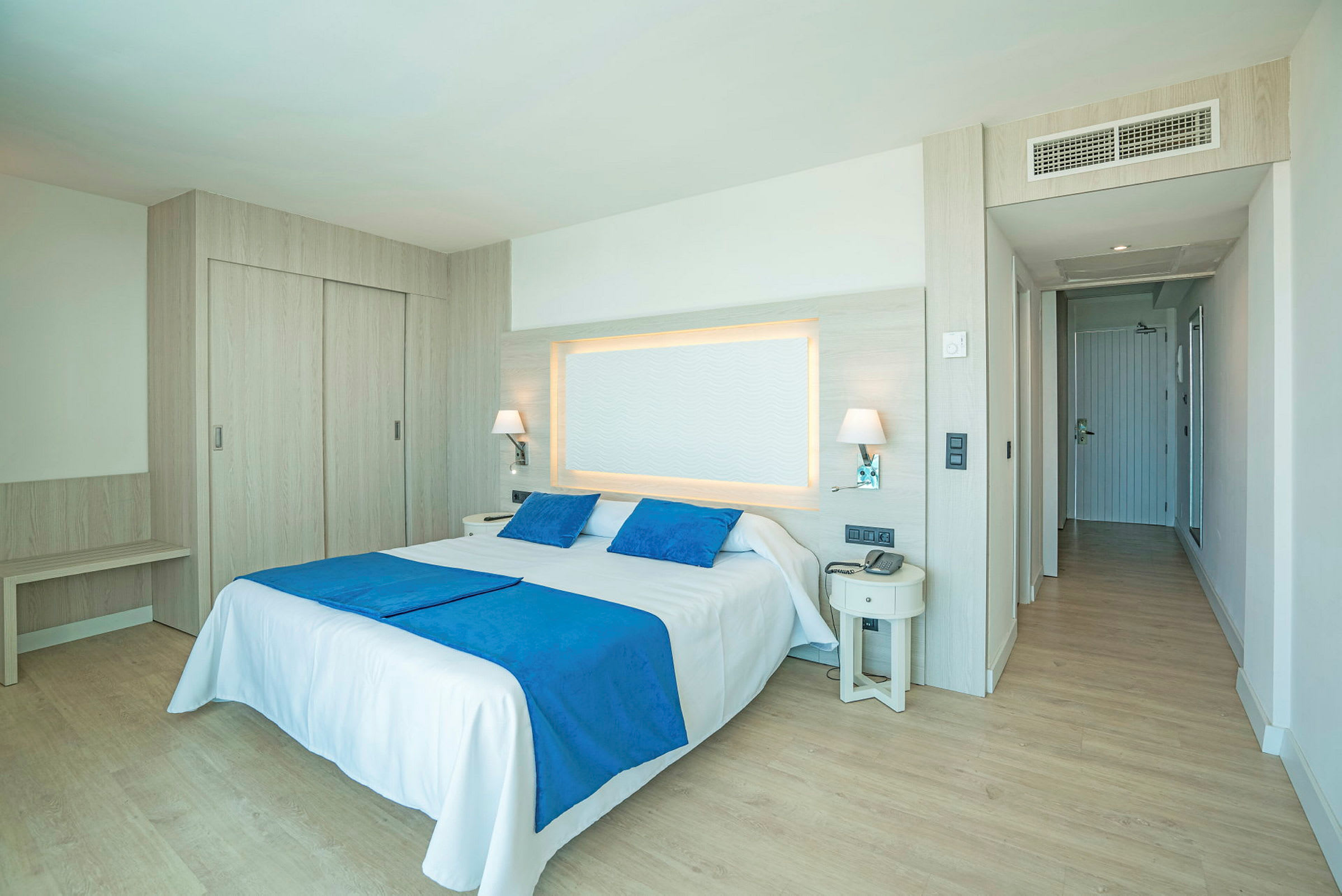 Ett hotellrum med en dubbelsäng, två blåa kuddar och ett blått överkast. Rummet har träpaneler och ett öppet utrymme. (AI-genererad text)