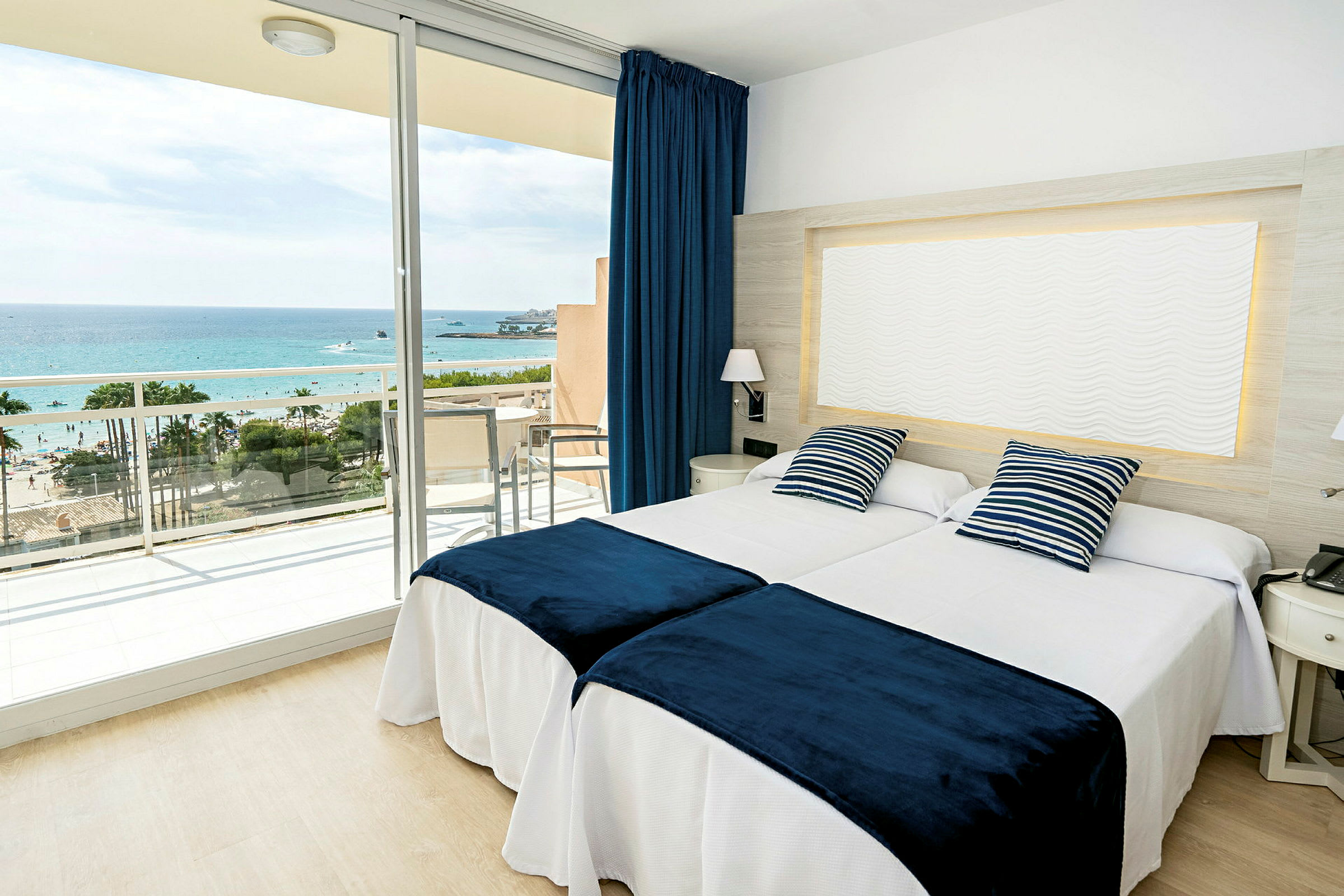 Ett hotellrum med två sängar, blåvita prydnadskuddar och blå överkast, samt en balkong med utsikt mot havet. (AI-genererad text)