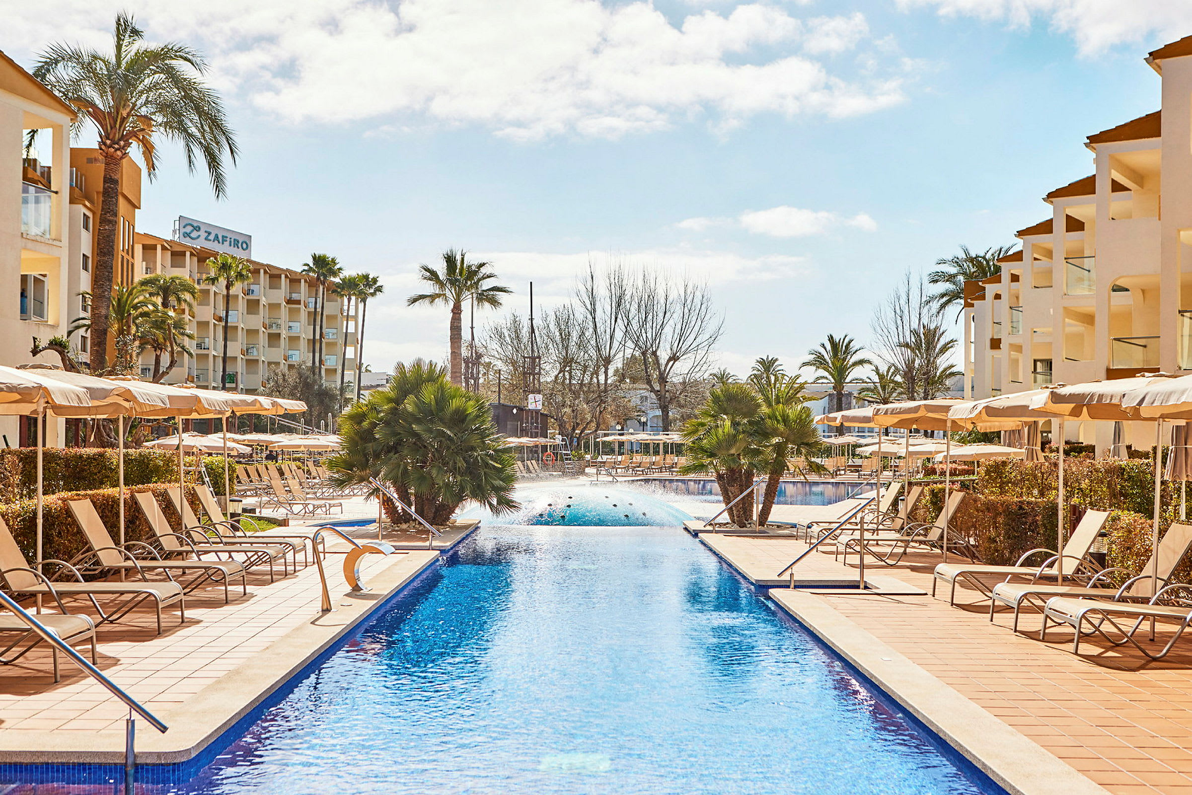 Bild av en lång swimmingpool omgiven av solstolar och parasoller, med hotellbyggnader i bakgrunden. (AI-genererad text)