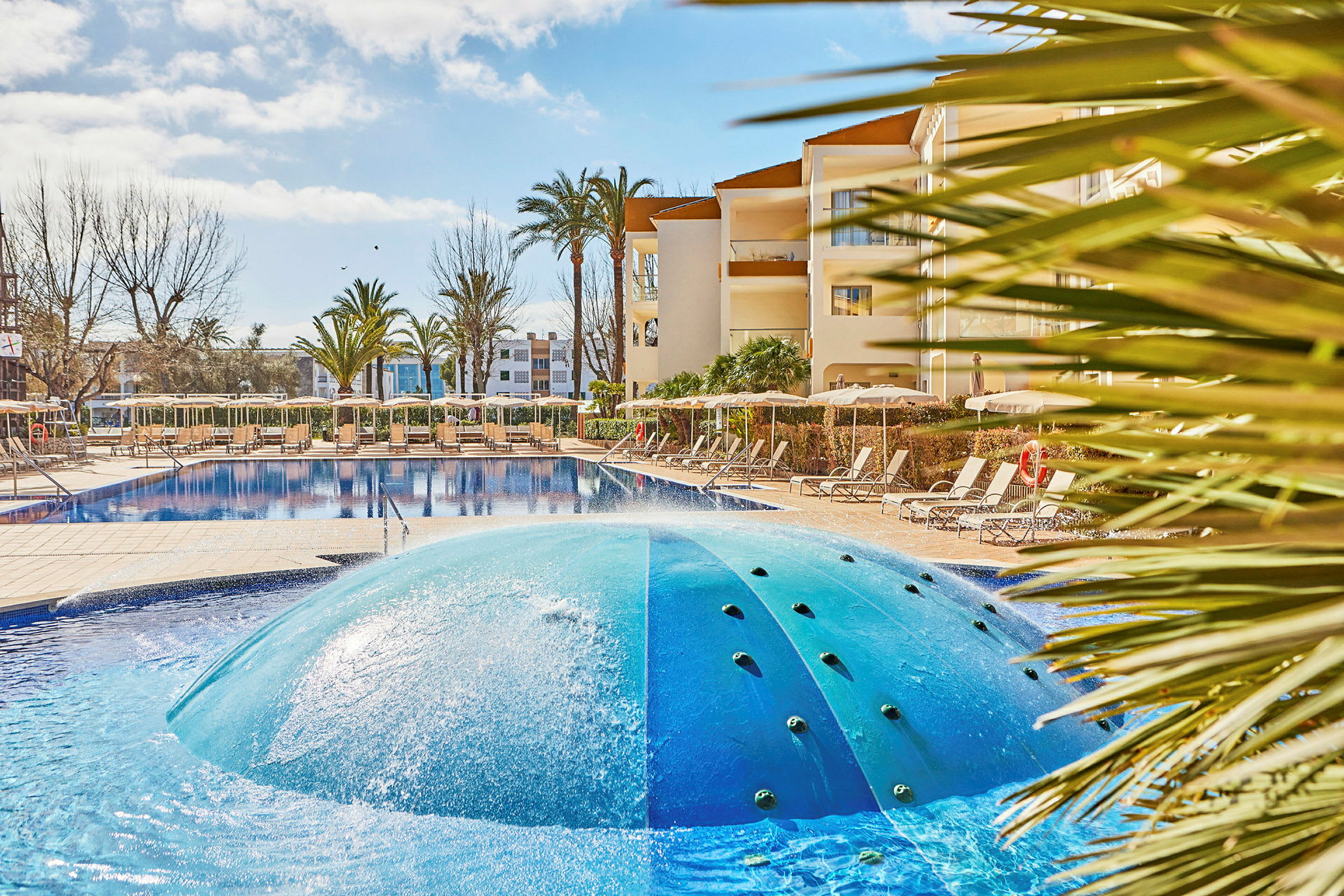 Bild av en pool med vågformad fontän i förgrunden, omgiven av solstolar och ett hotell i bakgrunden. (AI-genererad text)
