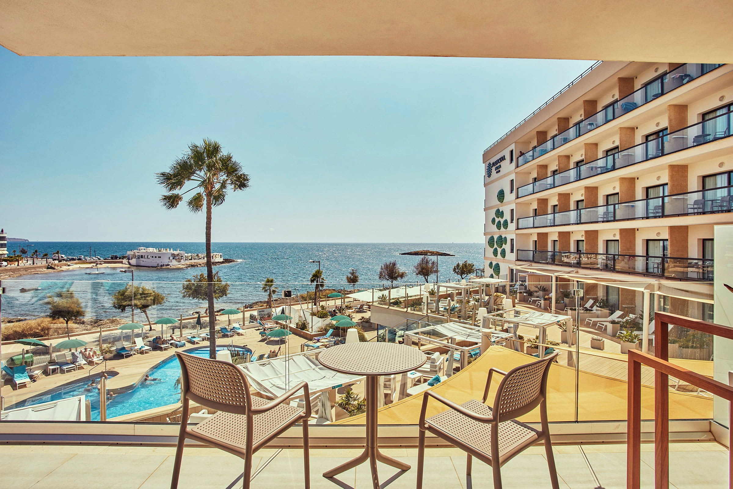 Bilden visar en balkong med två stolar och ett bord som vetter mot en pool och havsutsikt. Ett hotell med balkonger syns till höger. (AI-genererad text)