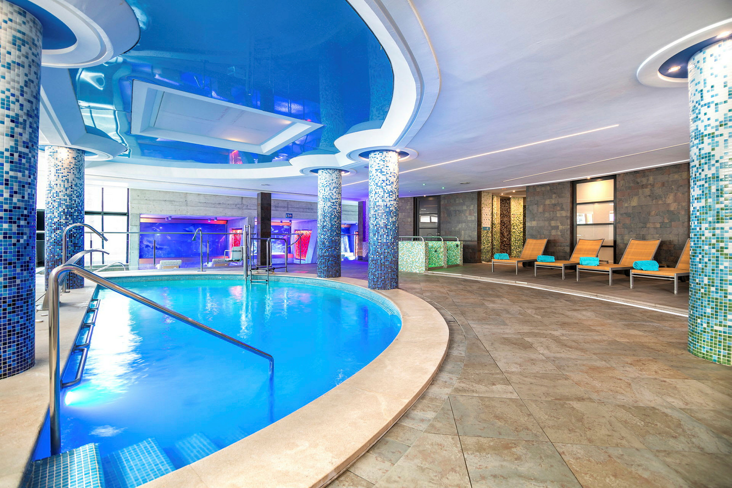 Inomhuspool i ett modernt spa med mosaikklädda pelare och vilstolar. (AI-genererad text)