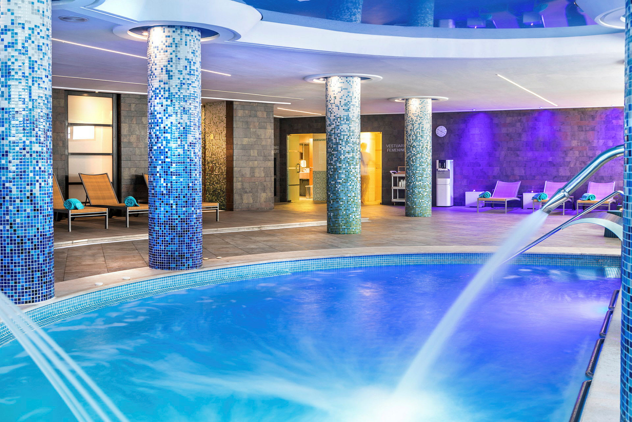 Inomhuspool med blå mosaik och fontän, omgiven av solstolar och väggar med en modern design. (AI-genererad text)