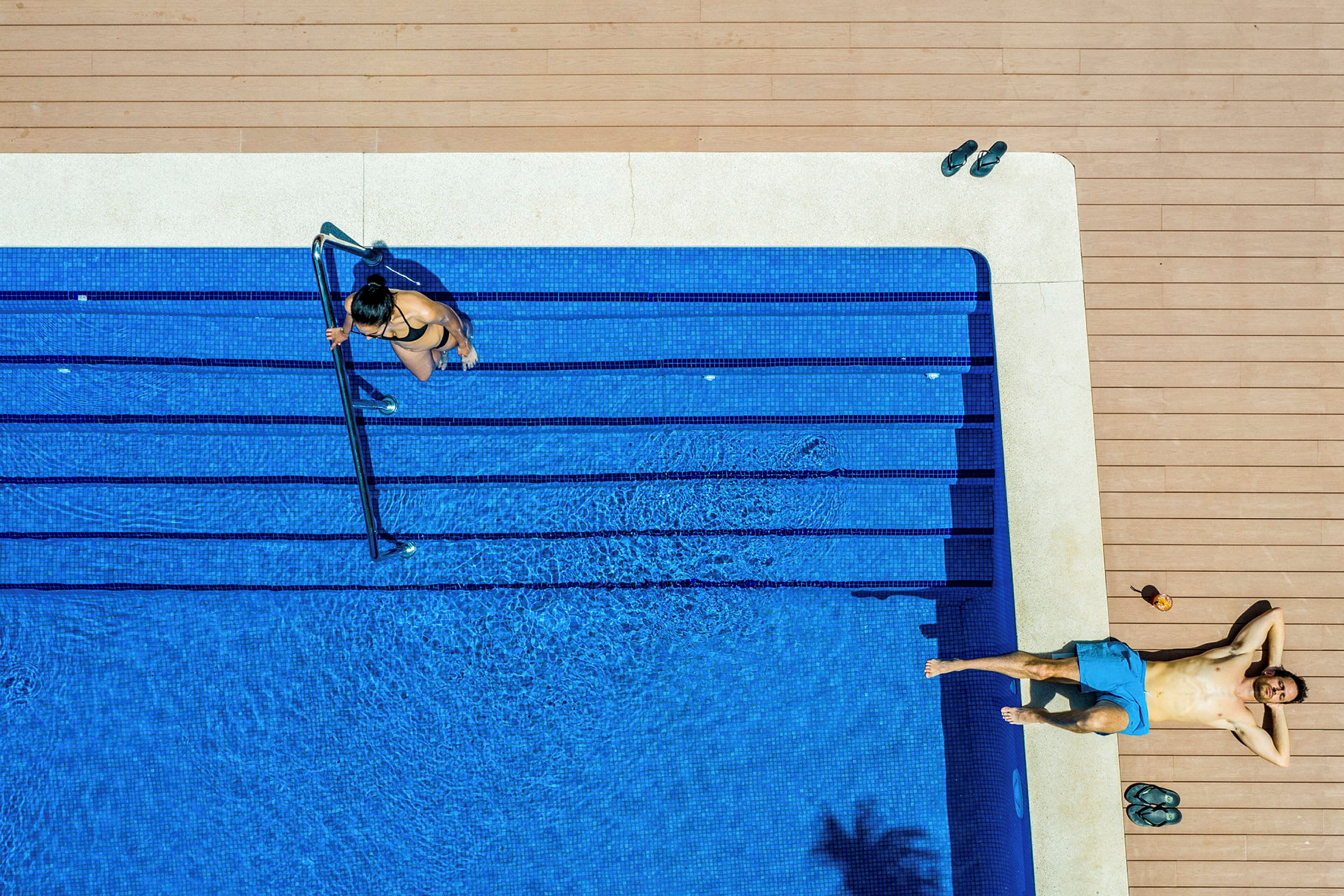 En kvinna går ner i en blå pool, och en man ligger på solsängen vid poolkanten. (AI-genererad text)