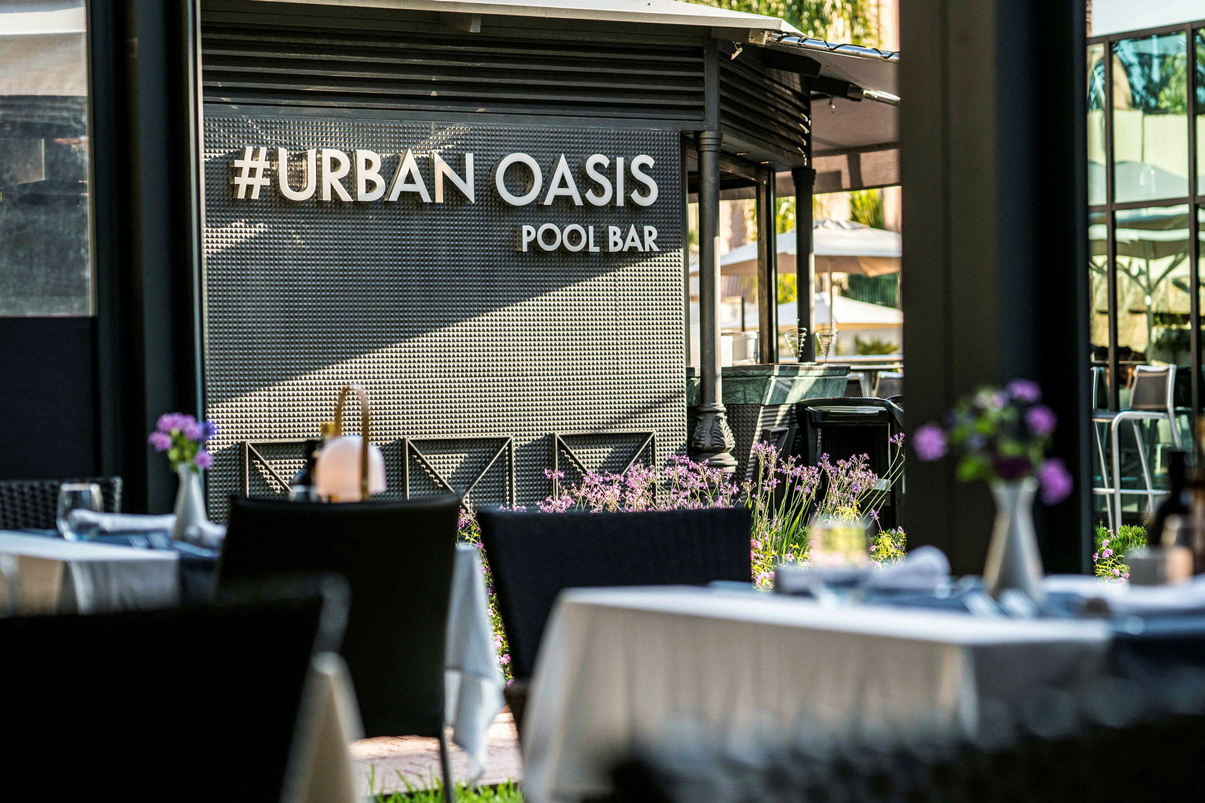 En poolbar med namnet #Urban Oasis, omgiven av blommor och utomhusbord. (AI-genererad text)