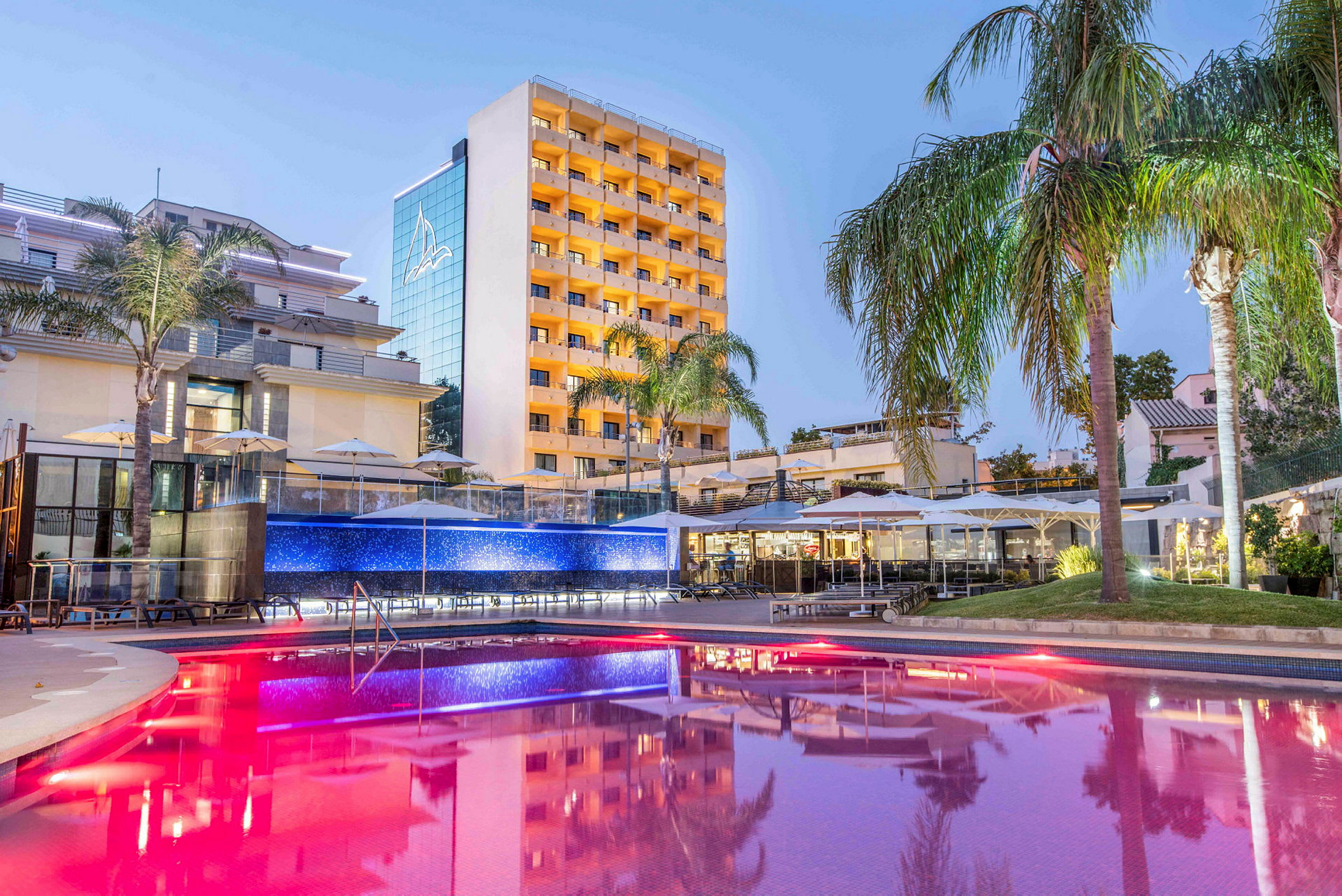 Hotellbyggnad med upplyst pool och palmer. (AI-genererad text)