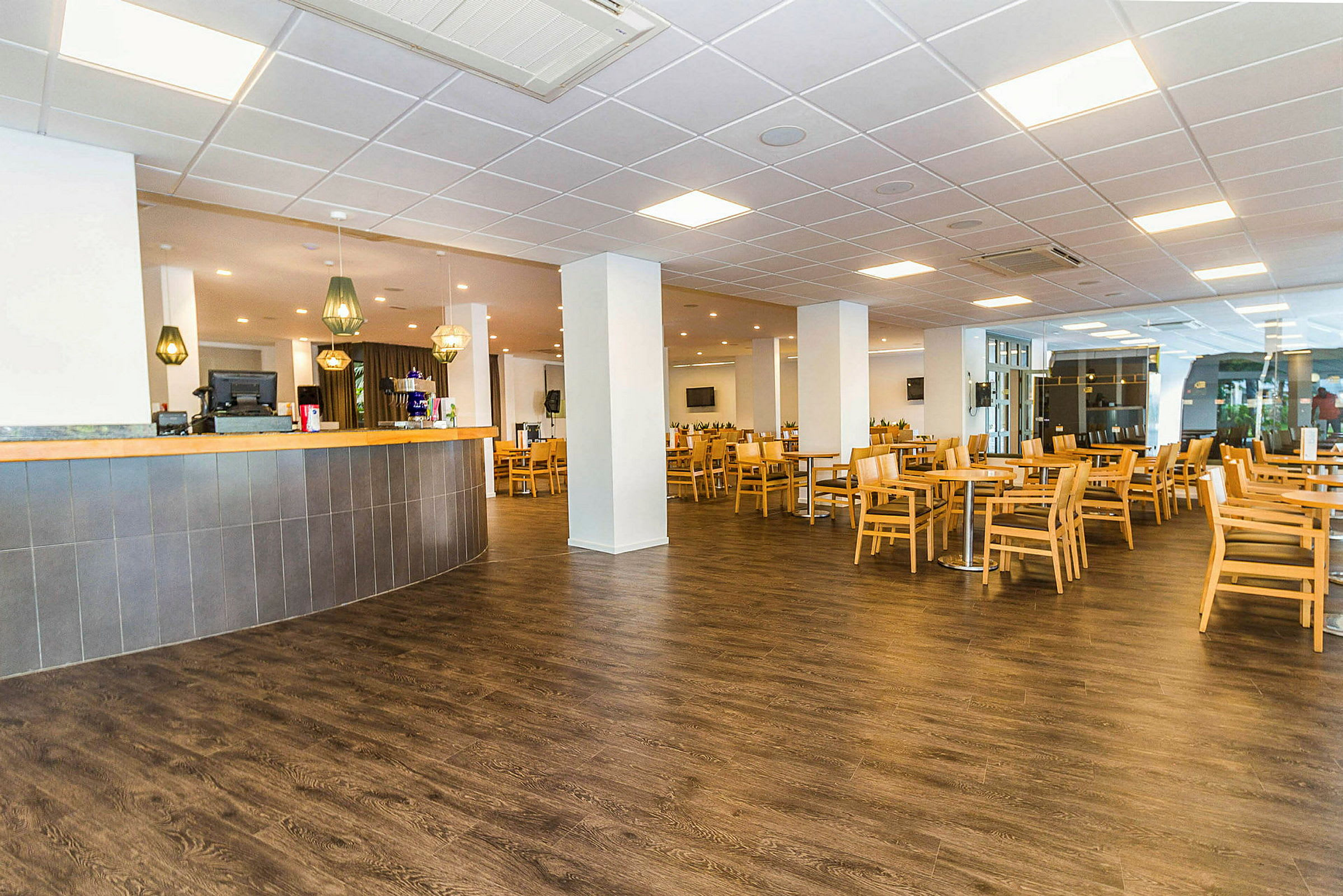 Ett rymligt café eller restaurangområde med ljusa bord och stolar i träplacerade över ett mörkt golv. I förgrunden finns en bar med hängande lampor ovanför. (AI-genererad text)